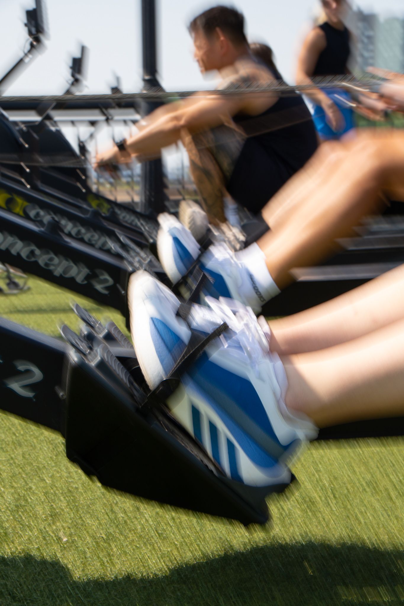 Close-up motion blur of athletes rowing on Concept2 machines on outdoor grass turf.