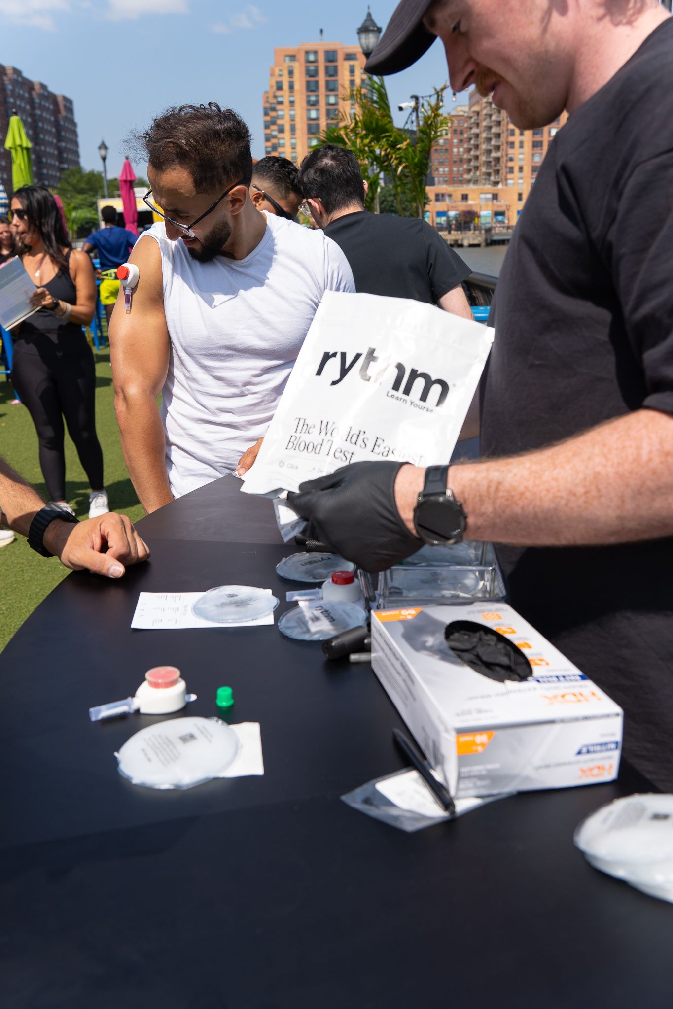 Two men examine Rytm wellness blood testing supplies at an outdoor health event on a sunny day.