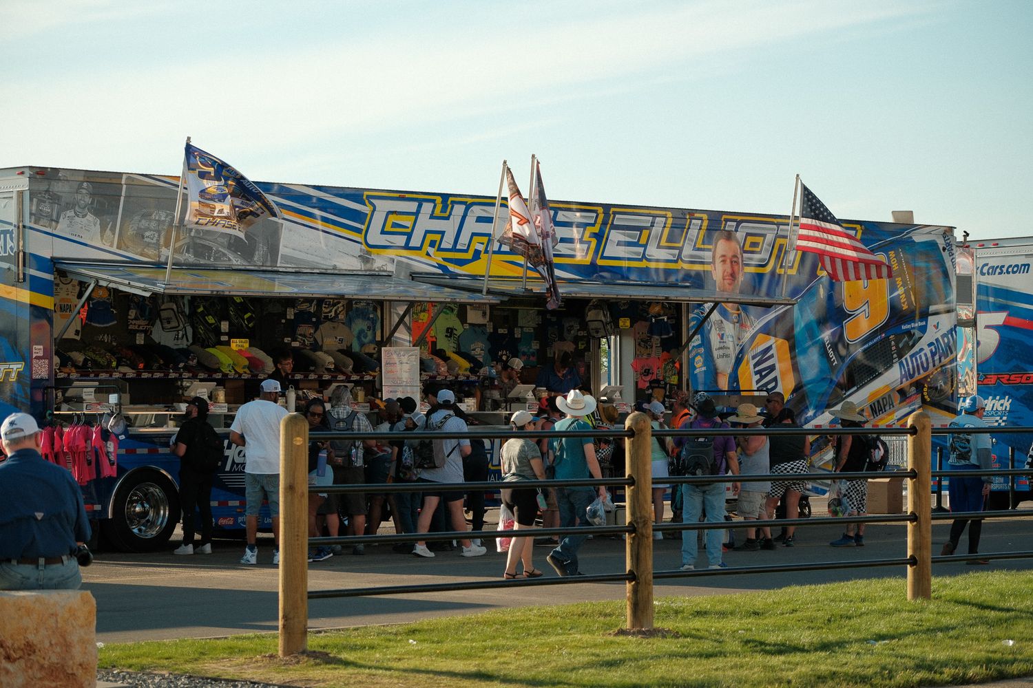 Fans browse a Chase Elliott NASCAR merchandise trailer at a speedway event on a sunny day.