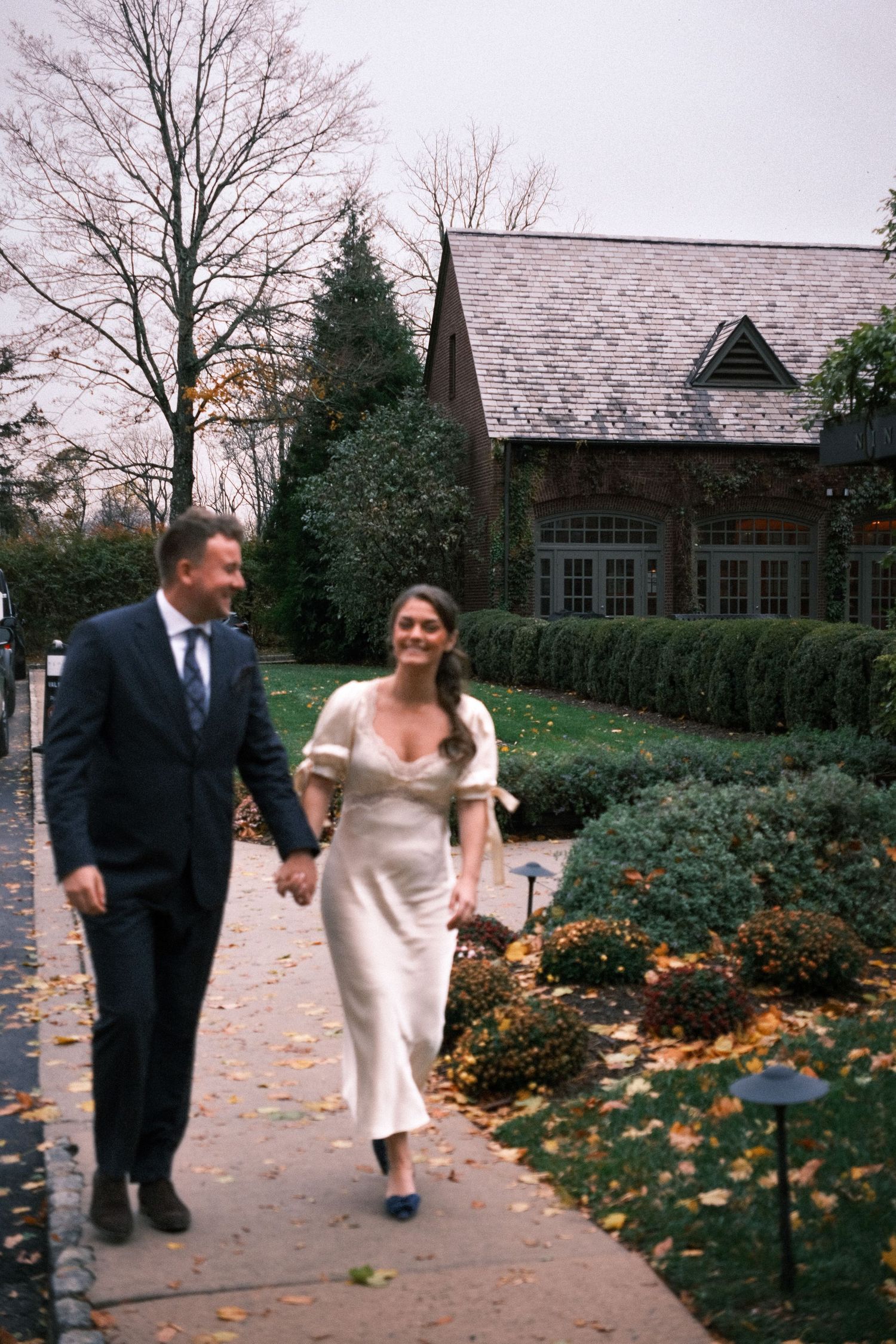 Couple in wedding attire walking hand-in-hand on a sidewalk outside an elegant estate in autumn.
