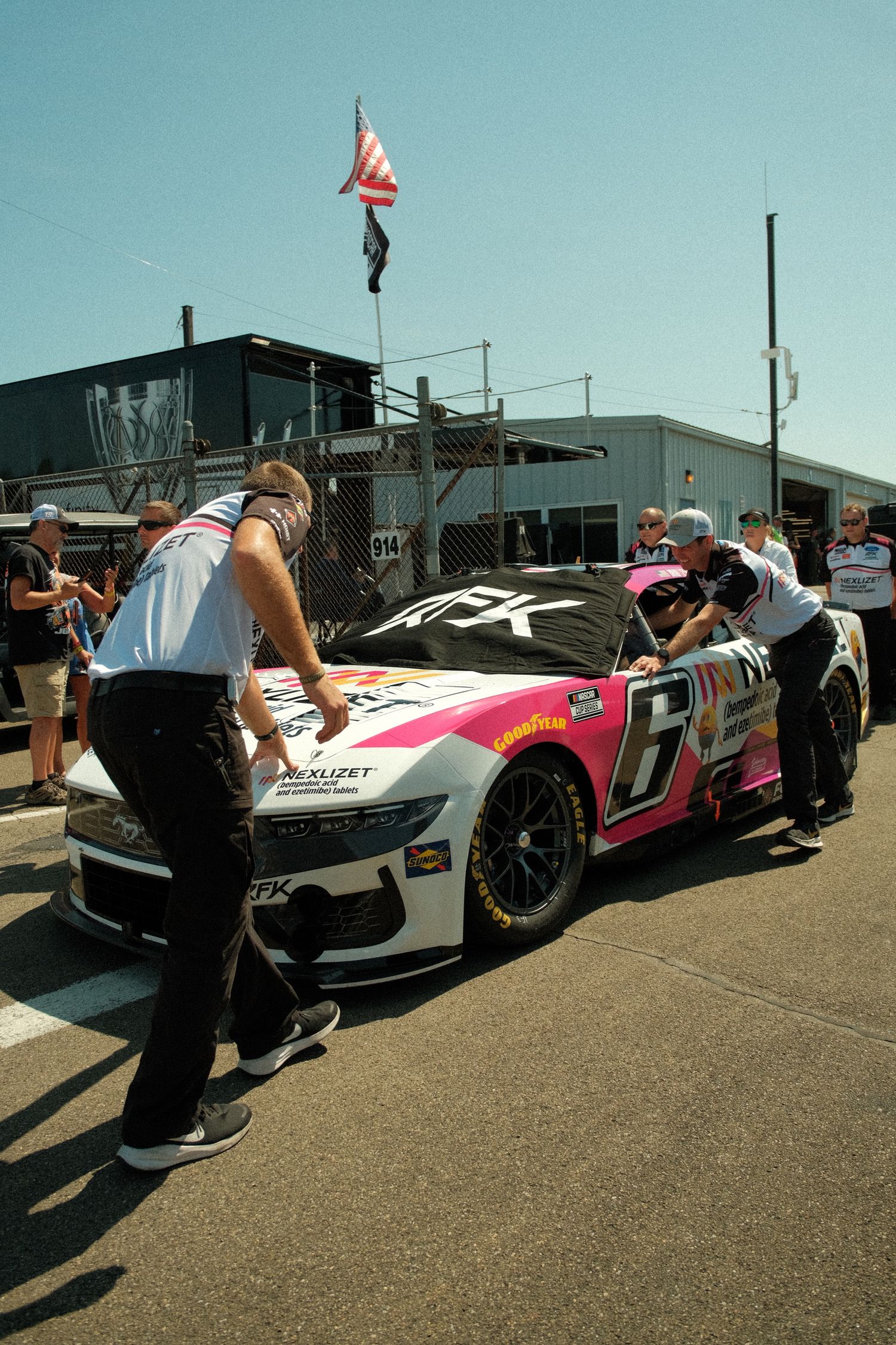 NASCAR pit crew members push the pink and white No. 6 race car in the pit lane at a sunny speedway.