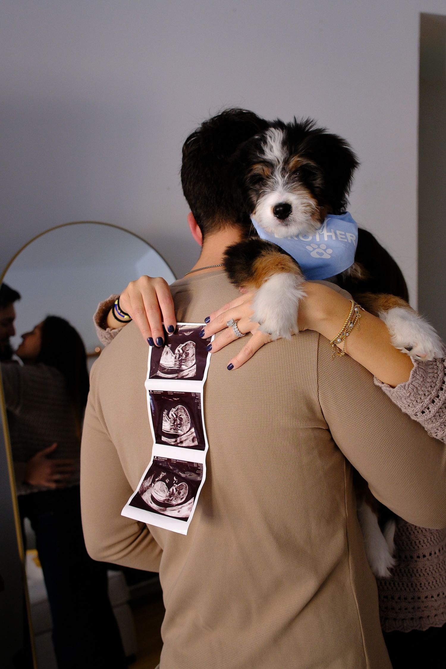 Couple embracing while holding ultrasound photos and a puppy wearing a bandana, announcing pregnancy.