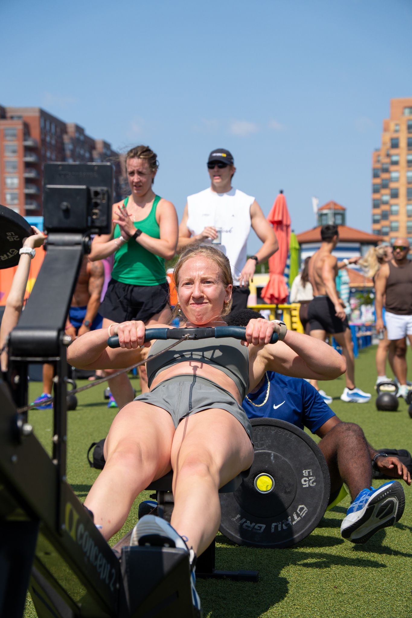 Woman performing barbell bench press exercise outdoors at a CrossFit competition on a sunny day.