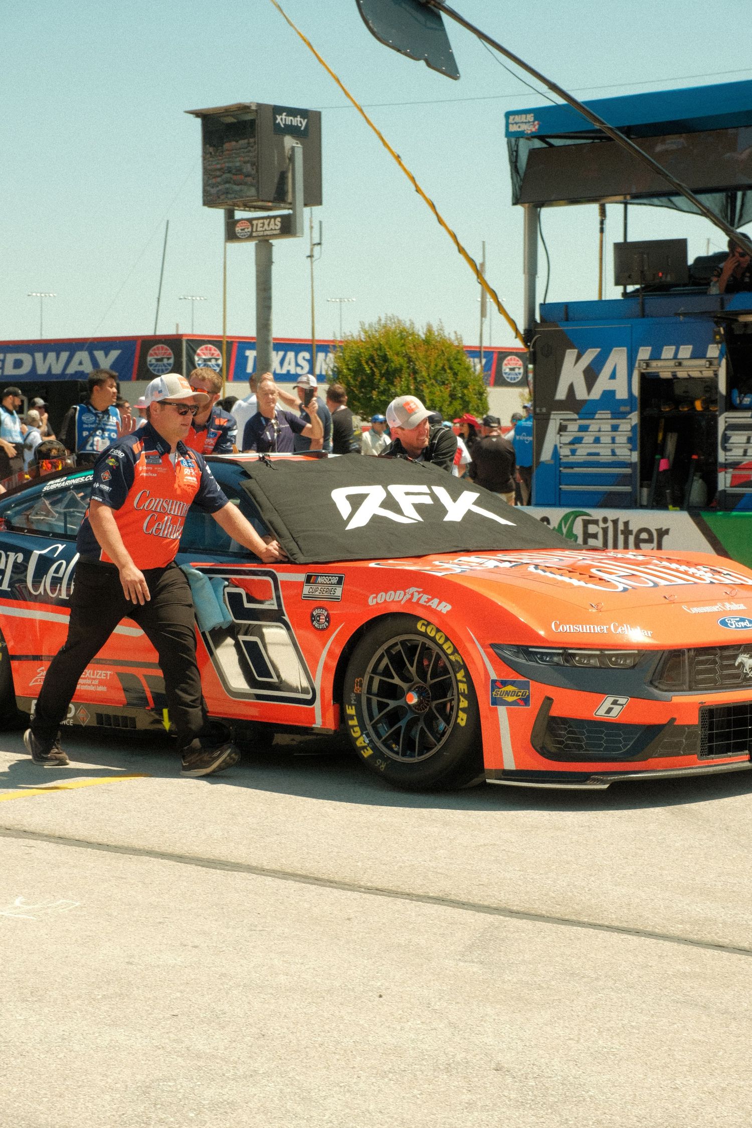 RFK Racing orange NASCAR Ford Mustang #6 being pushed by pit crew at Texas Motor Speedway.