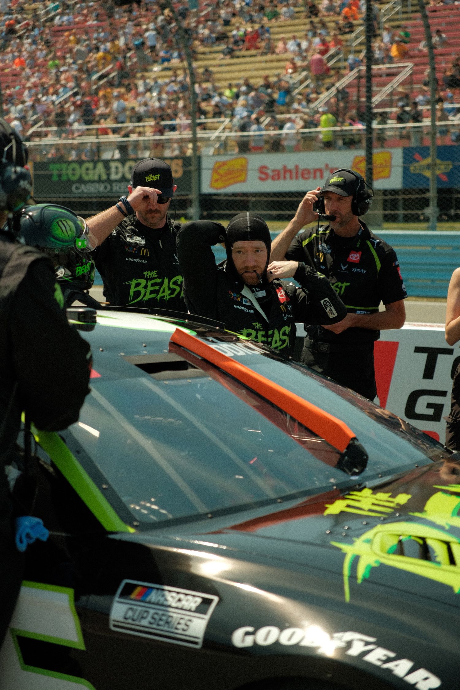 NASCAR Cup Series pit crew members working on black The Beast race car during race event at packed speedway.