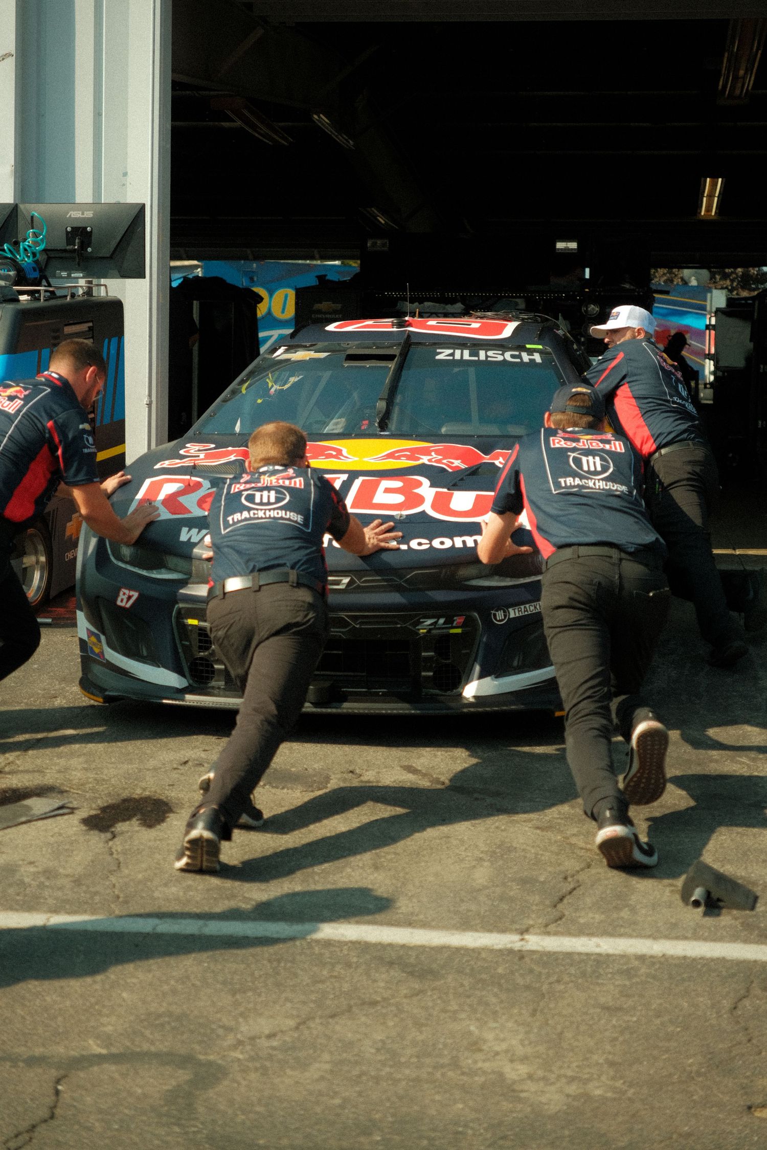Red Bull NASCAR pit crew members pushing dark race car out of garage stall.