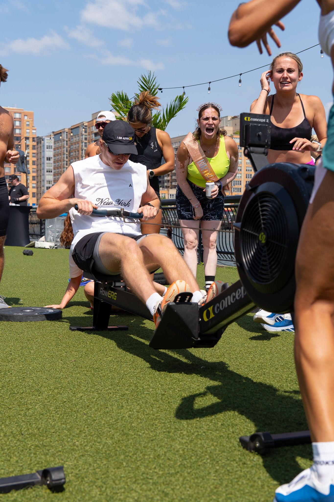 Group fitness class on rooftop turf, man rowing on Concept2 machine while cheering crowd watches on sunny day.
