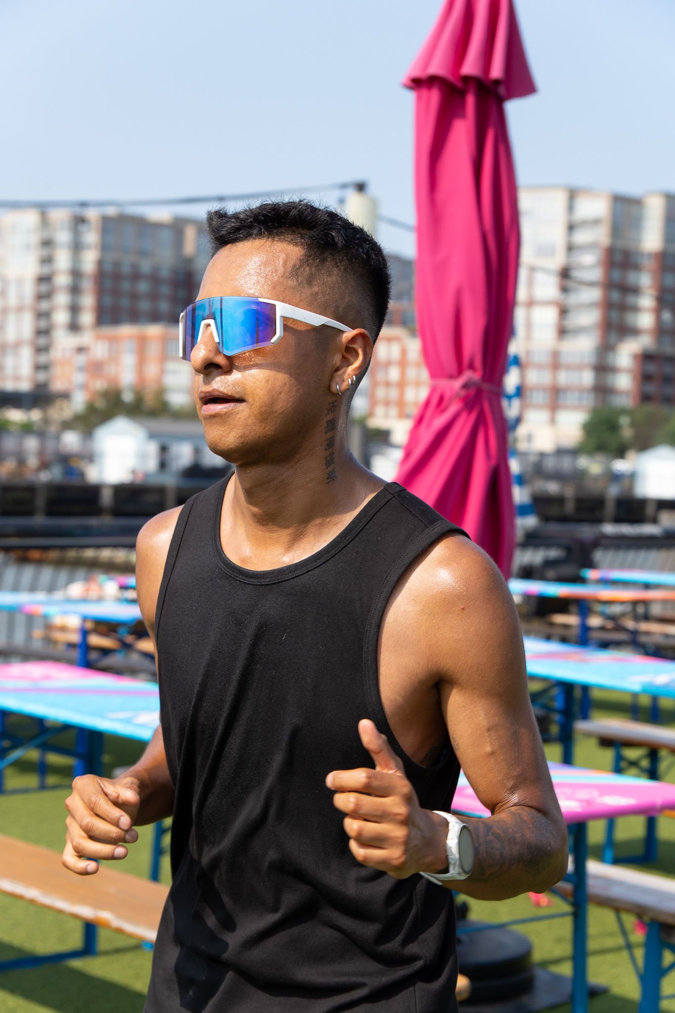 Athletic man in black tank top and blue mirrored sunglasses jogging outdoors at an urban venue on a sunny day.