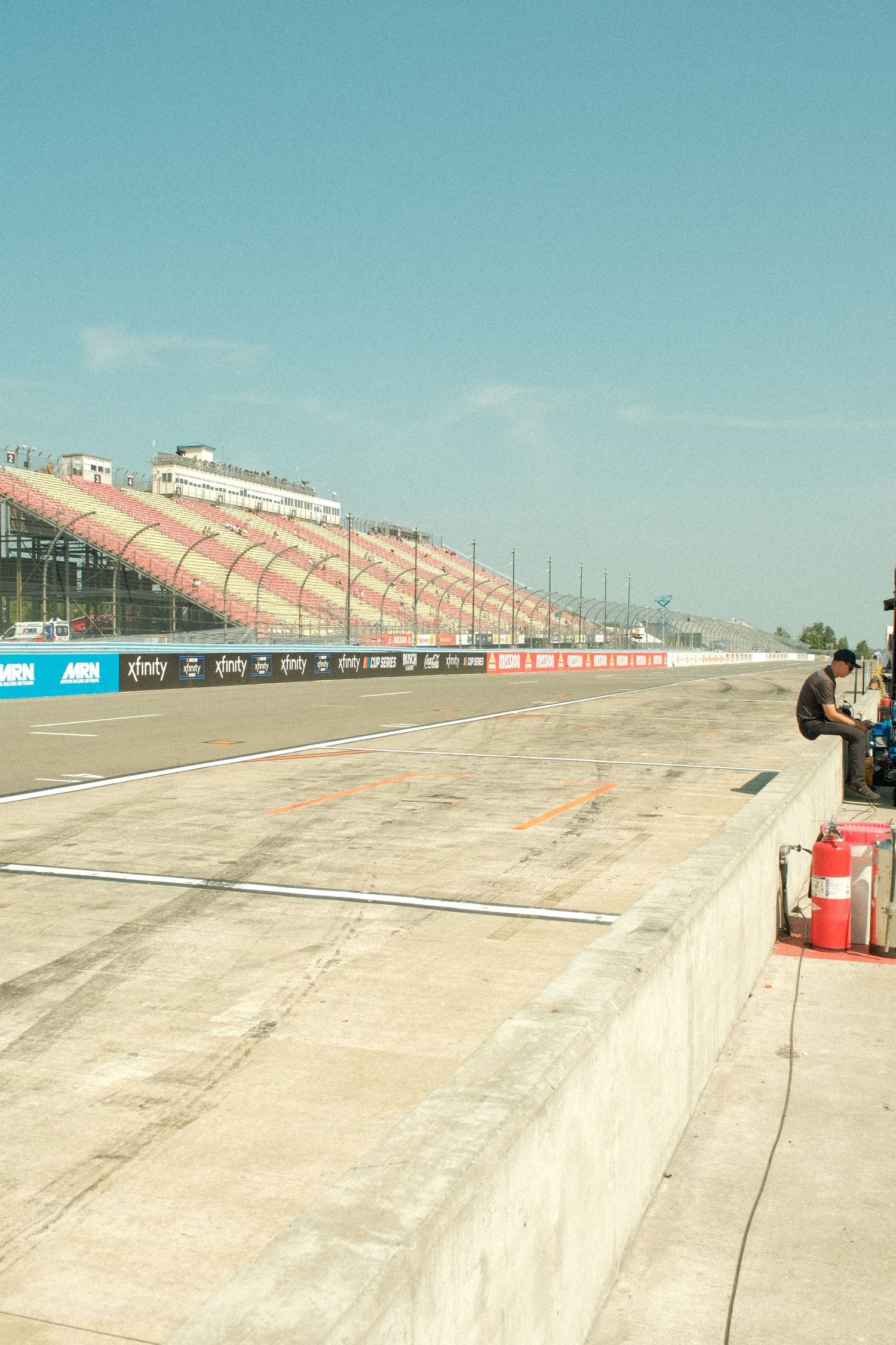 Empty NASCAR pit road with red and white grandstands visible under clear blue sky.