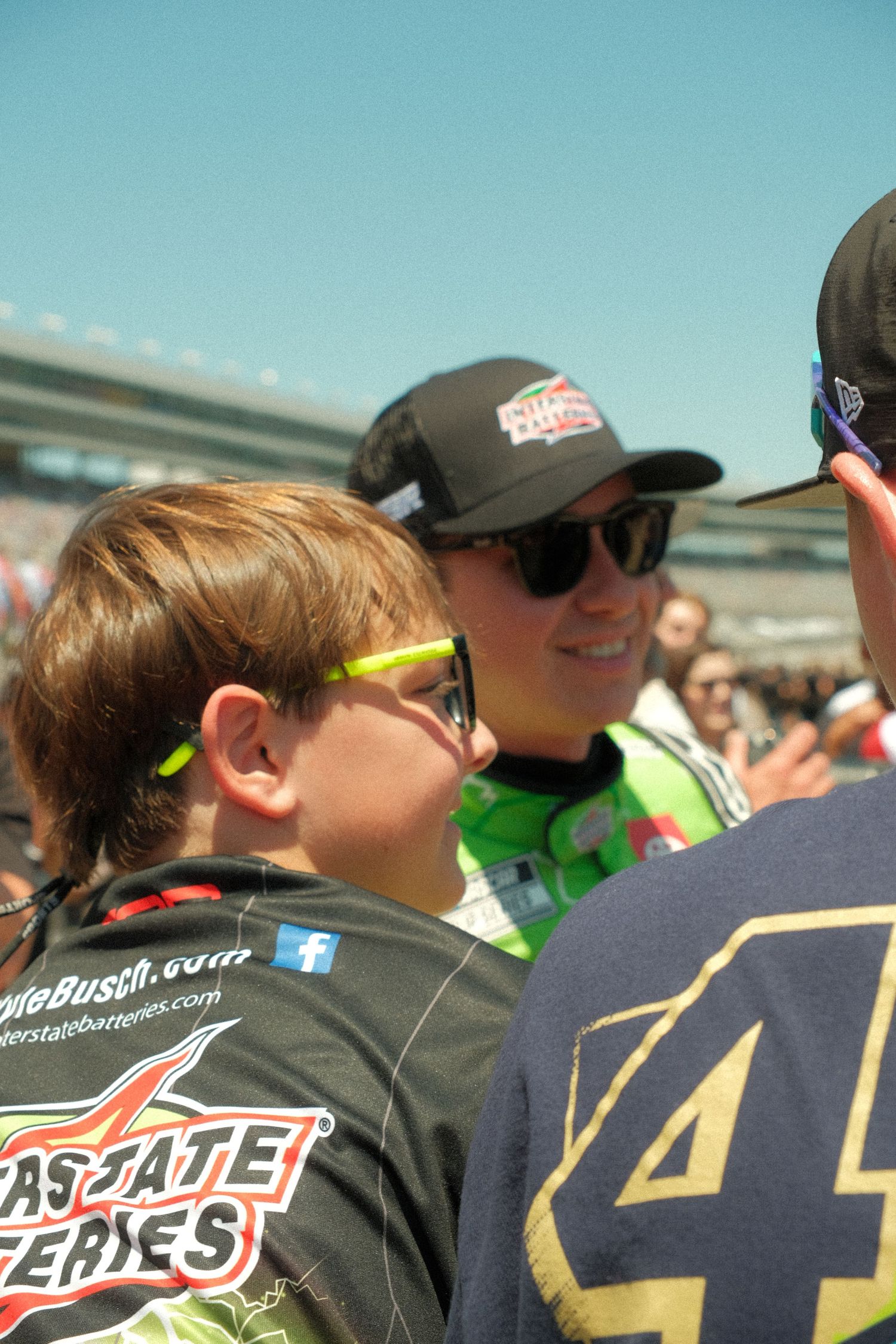 A young NASCAR fan wearing Interstate Batteries gear and yellow sunglasses at a sunny race event.