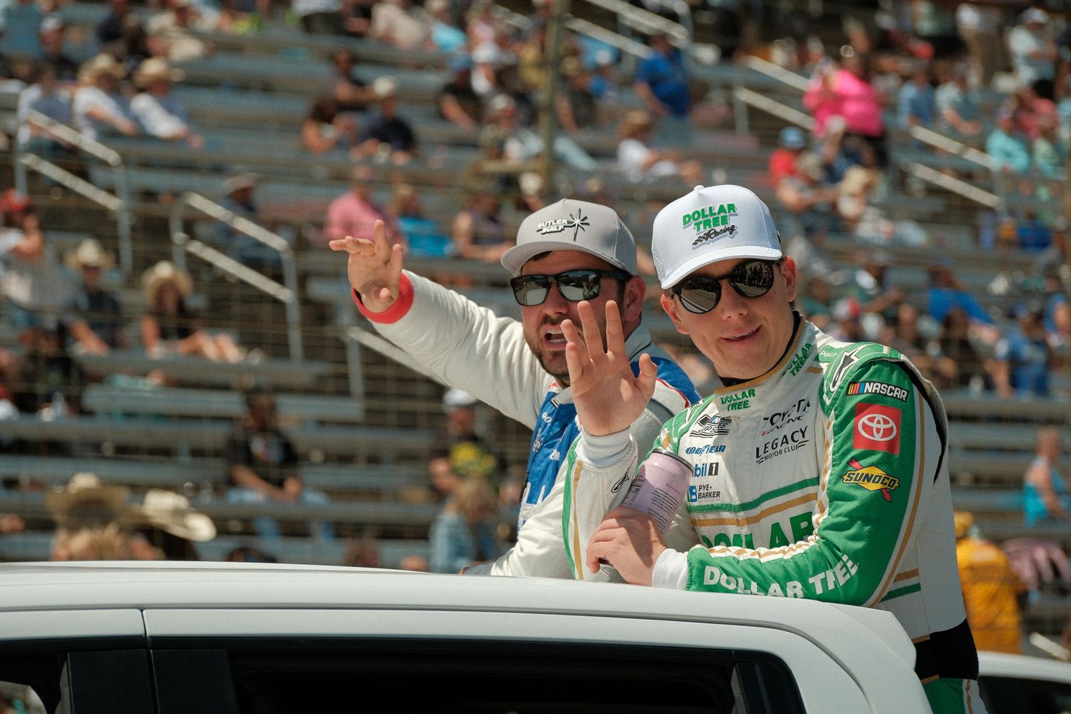 Two NASCAR drivers wave to fans during a pre-race driver parade at a packed speedway.