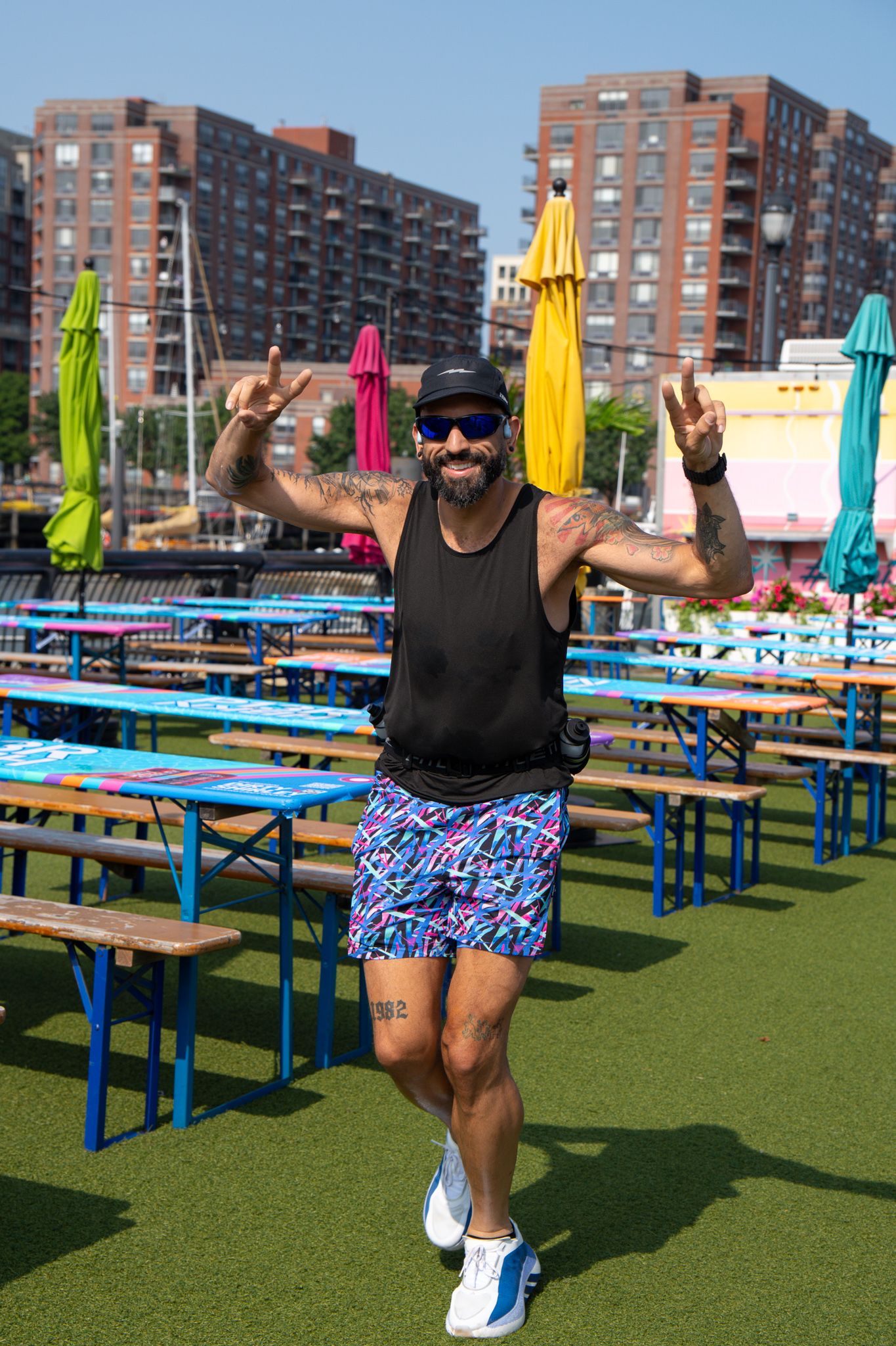 Energetic tattooed man posing with peace signs at colorful outdoor beer garden near urban waterfront.