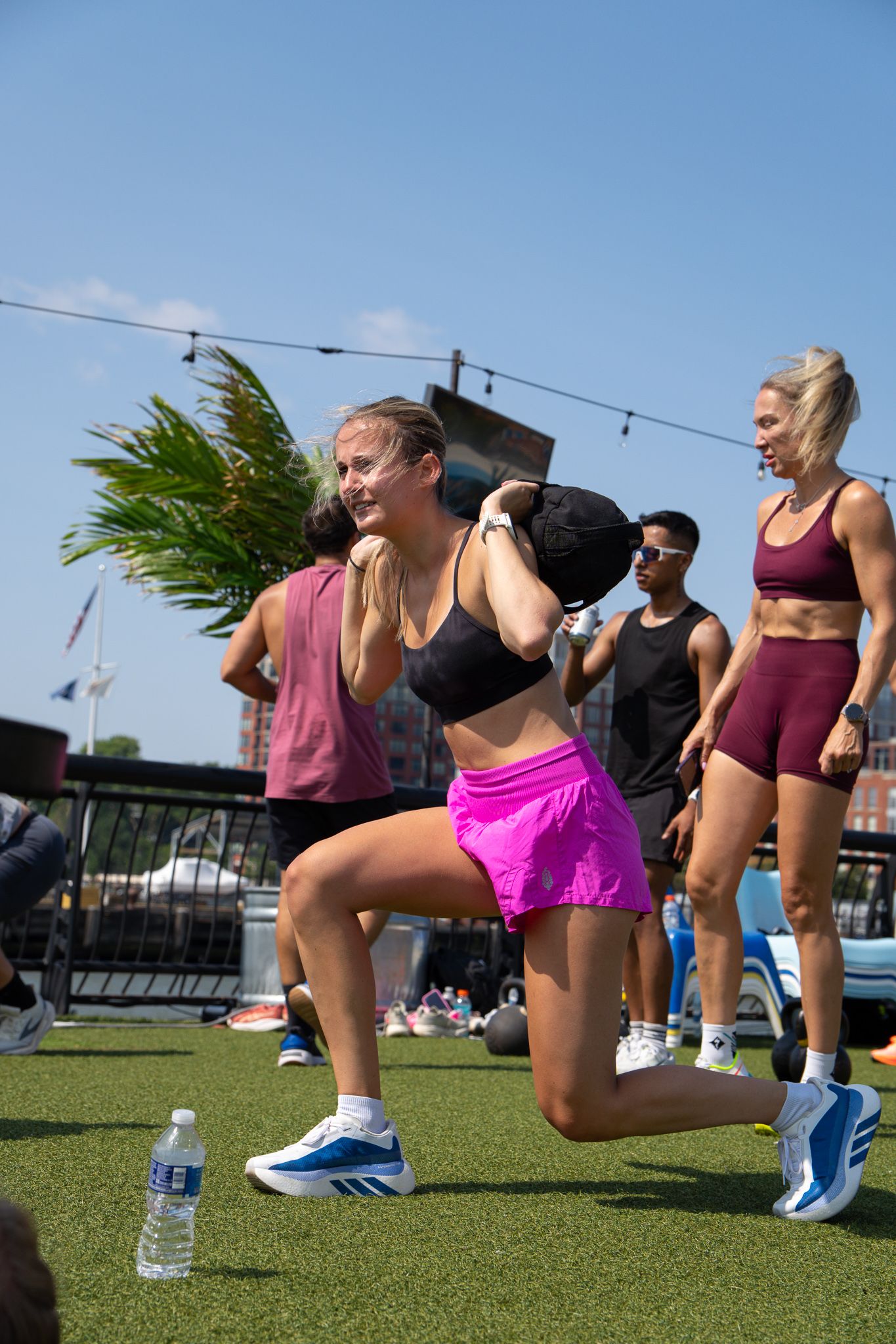 Athletic woman doing lunges with sandbag during outdoor group fitness class on artificial turf.