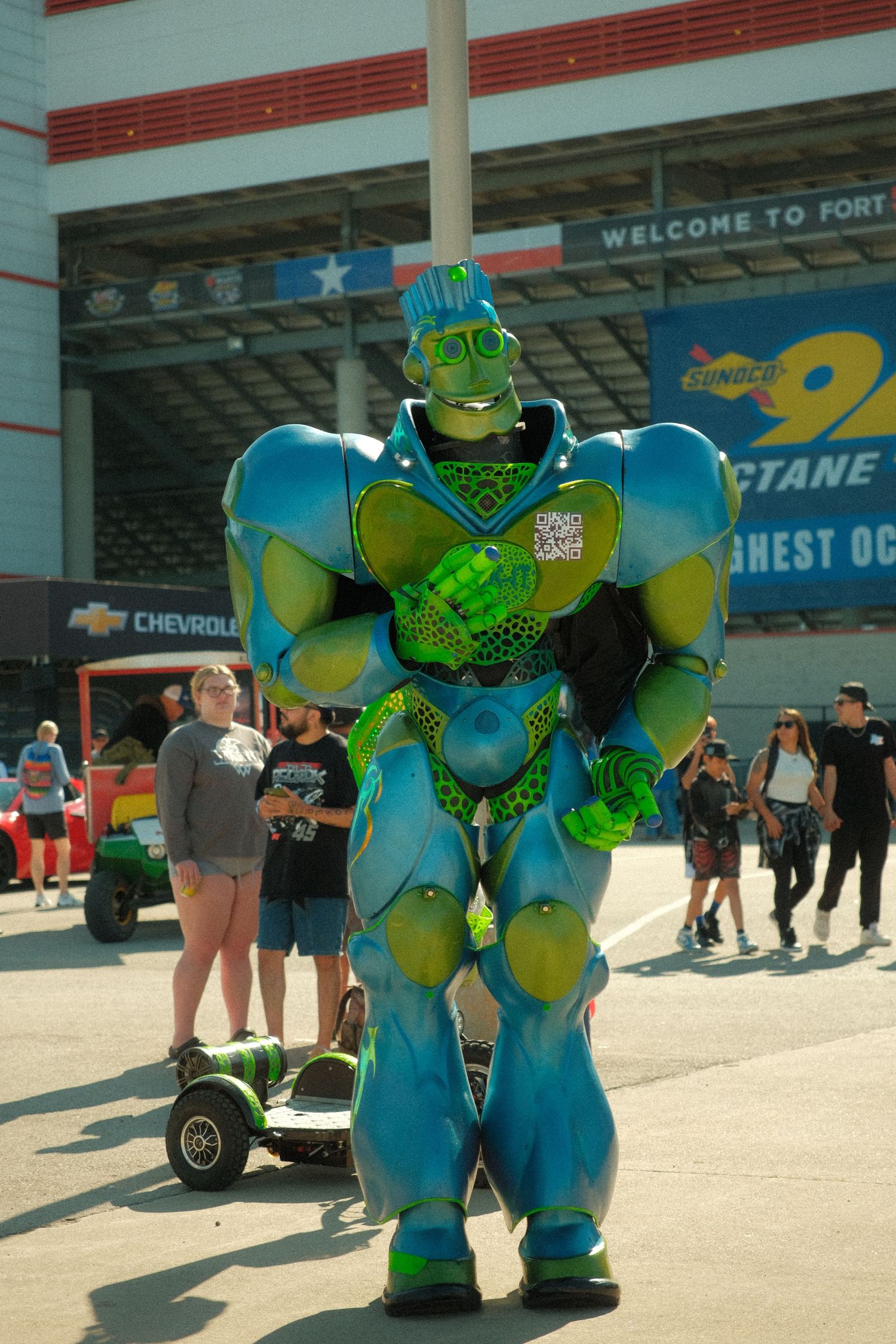 Colorful blue and green robot mascot costume entertains fans at Fort Worth NASCAR race event.