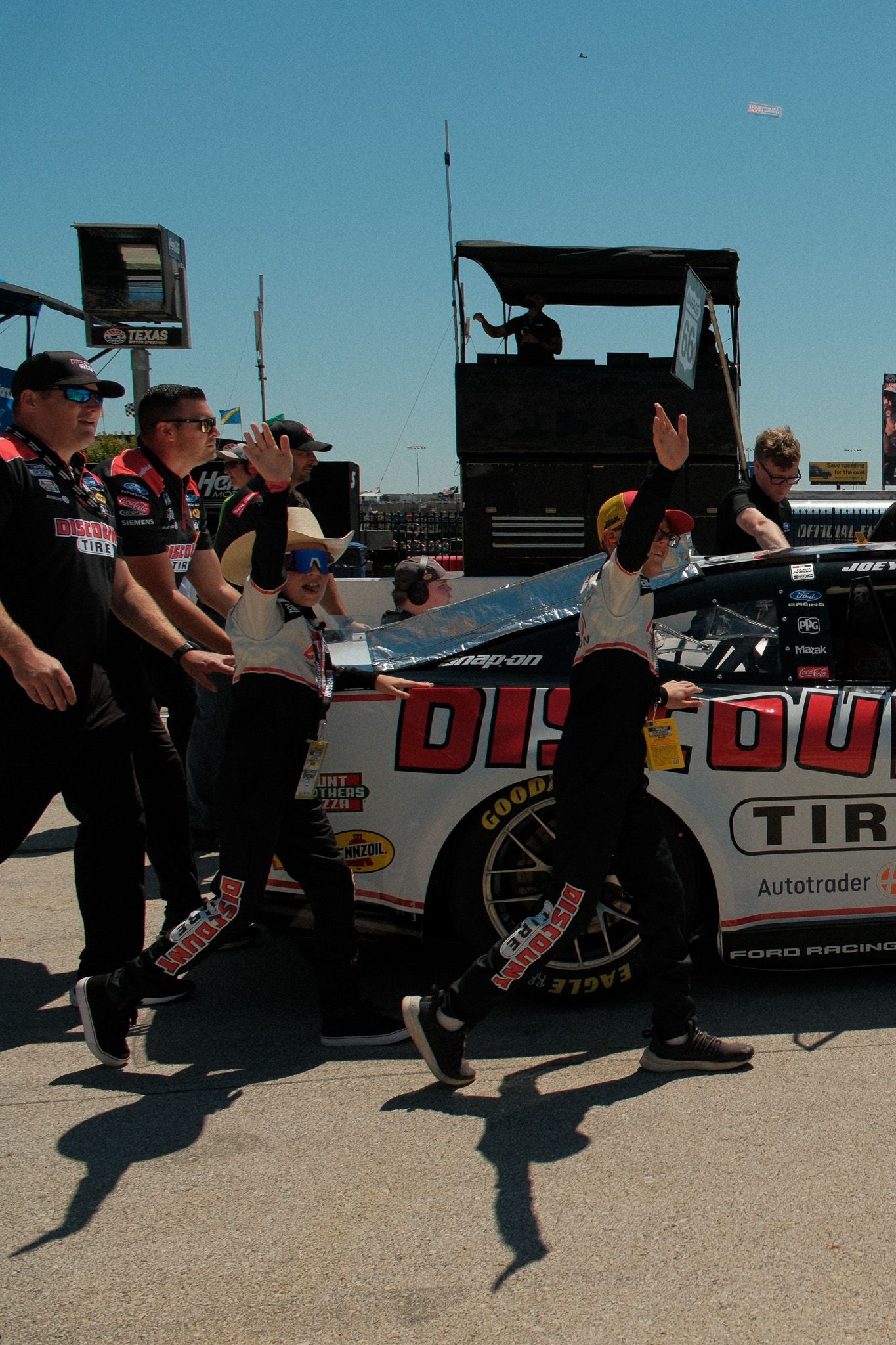 Discount Tire NASCAR pit crew pushes Ford Racing car through paddock area in sunlight.