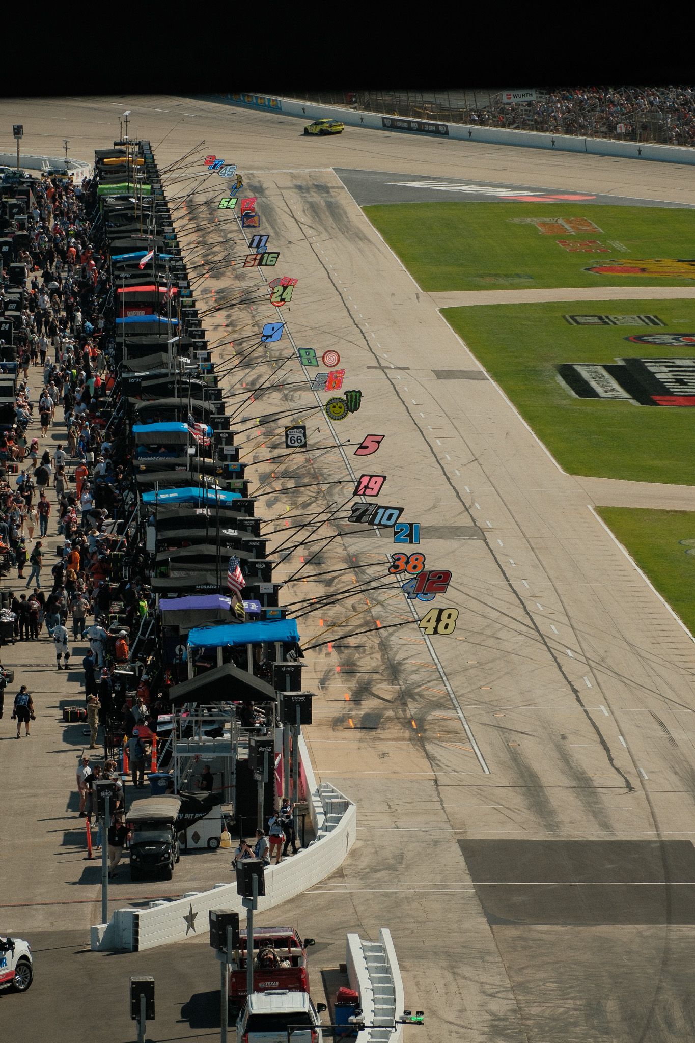 Aerial view of NASCAR pit road with numbered stalls, crew members, and race cars lined up along the track.