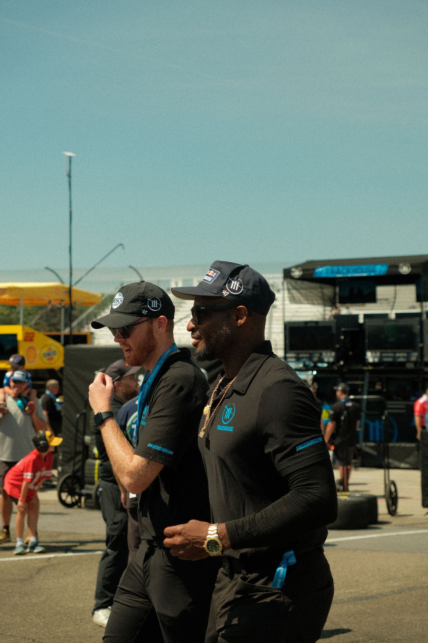 Two NASCAR team members in matching black uniforms walk through busy speedway paddock on sunny race day.
