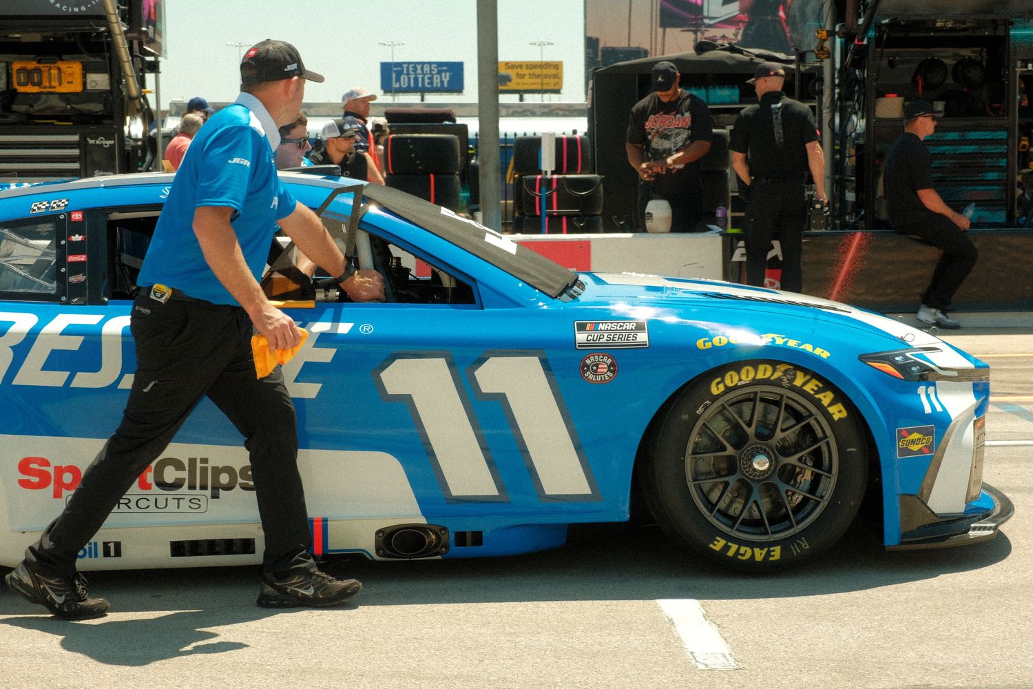 NASCAR #11 blue race car being pushed through pit road by crew members at a sunny speedway event.