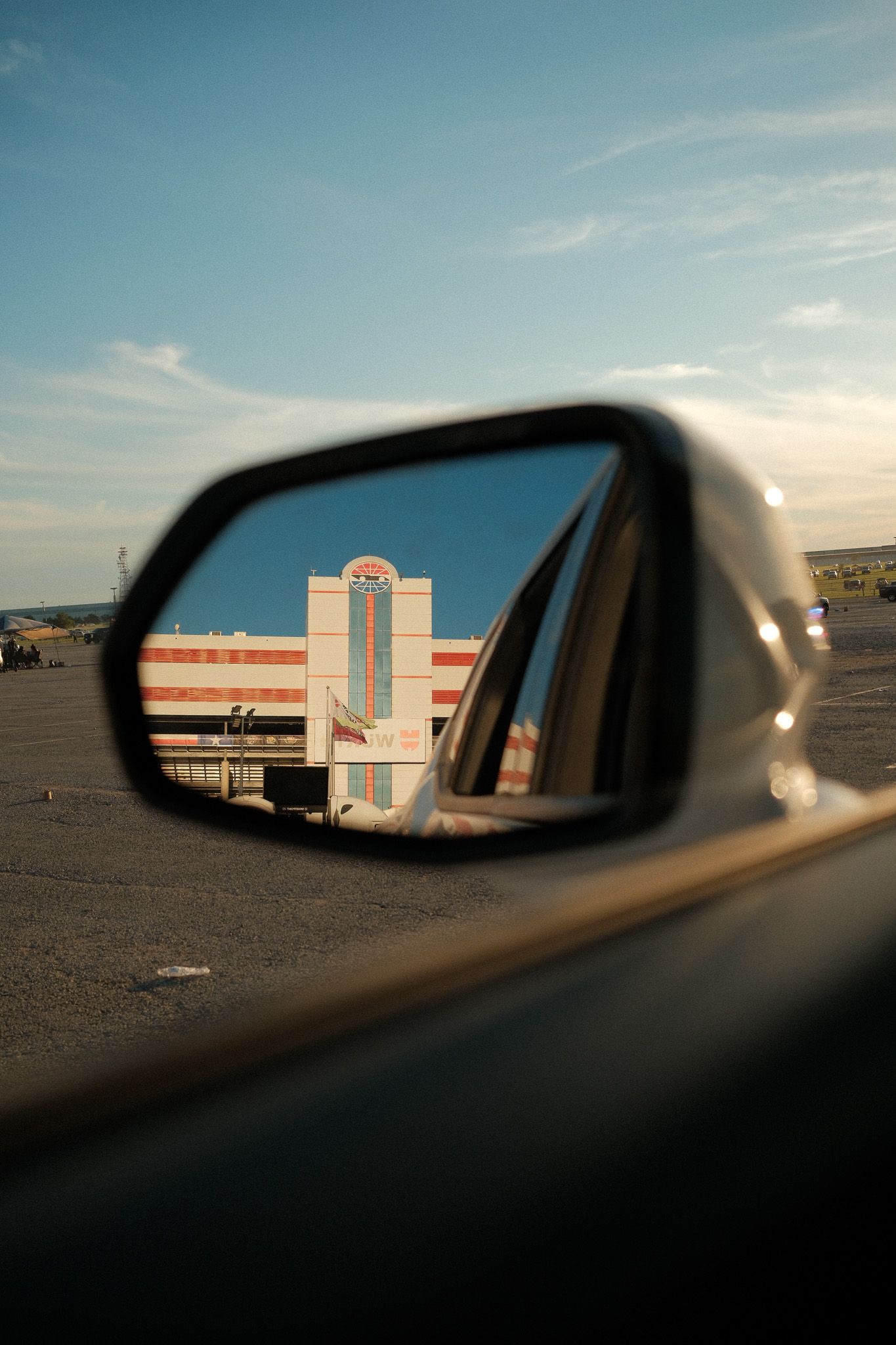 A car side mirror reflects a colorful building facade against a blue sky in a parking lot.