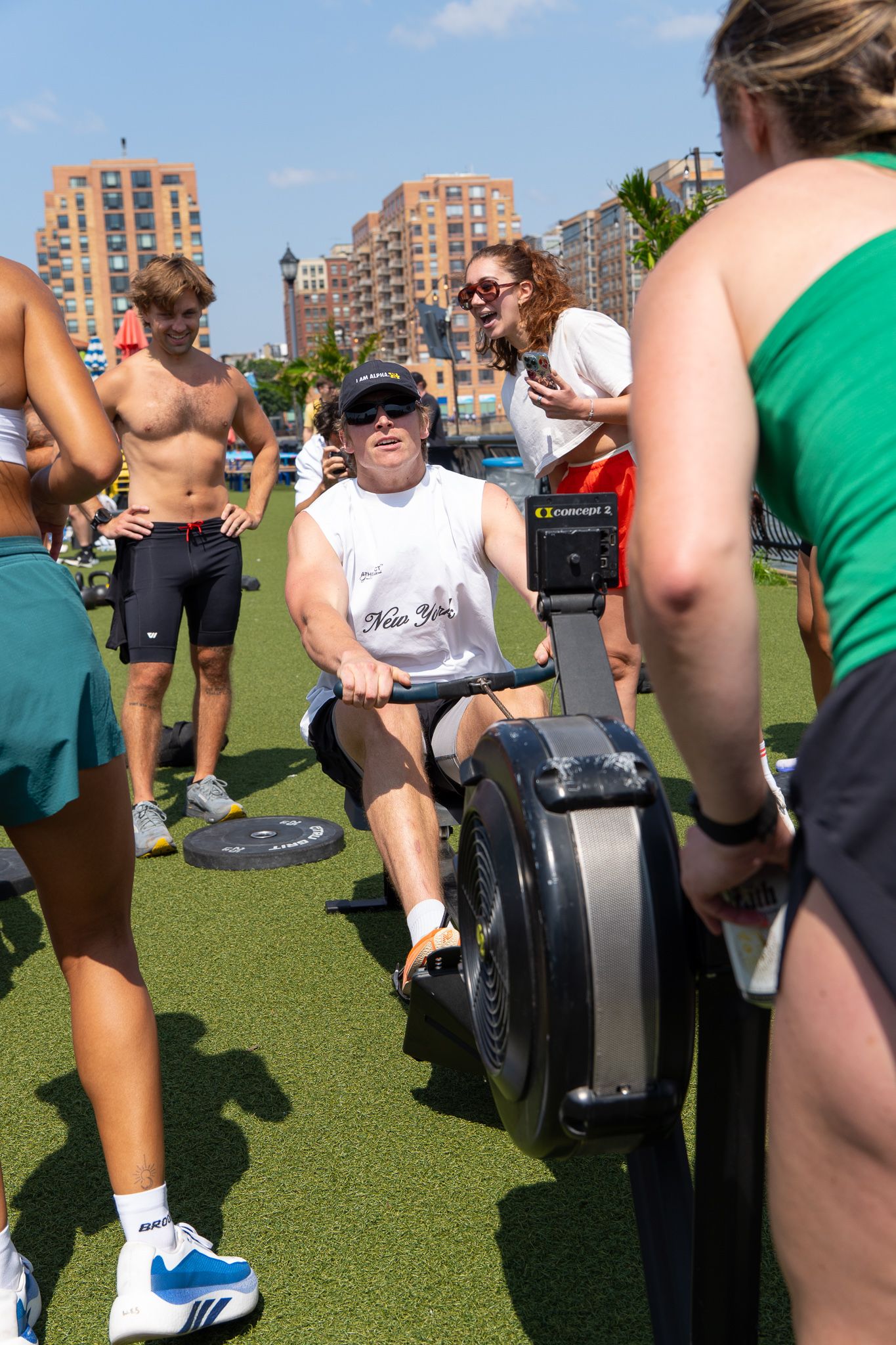 Man rowing on an ergometer machine outdoors on turf, surrounded by spectators, with NYC buildings in the background.