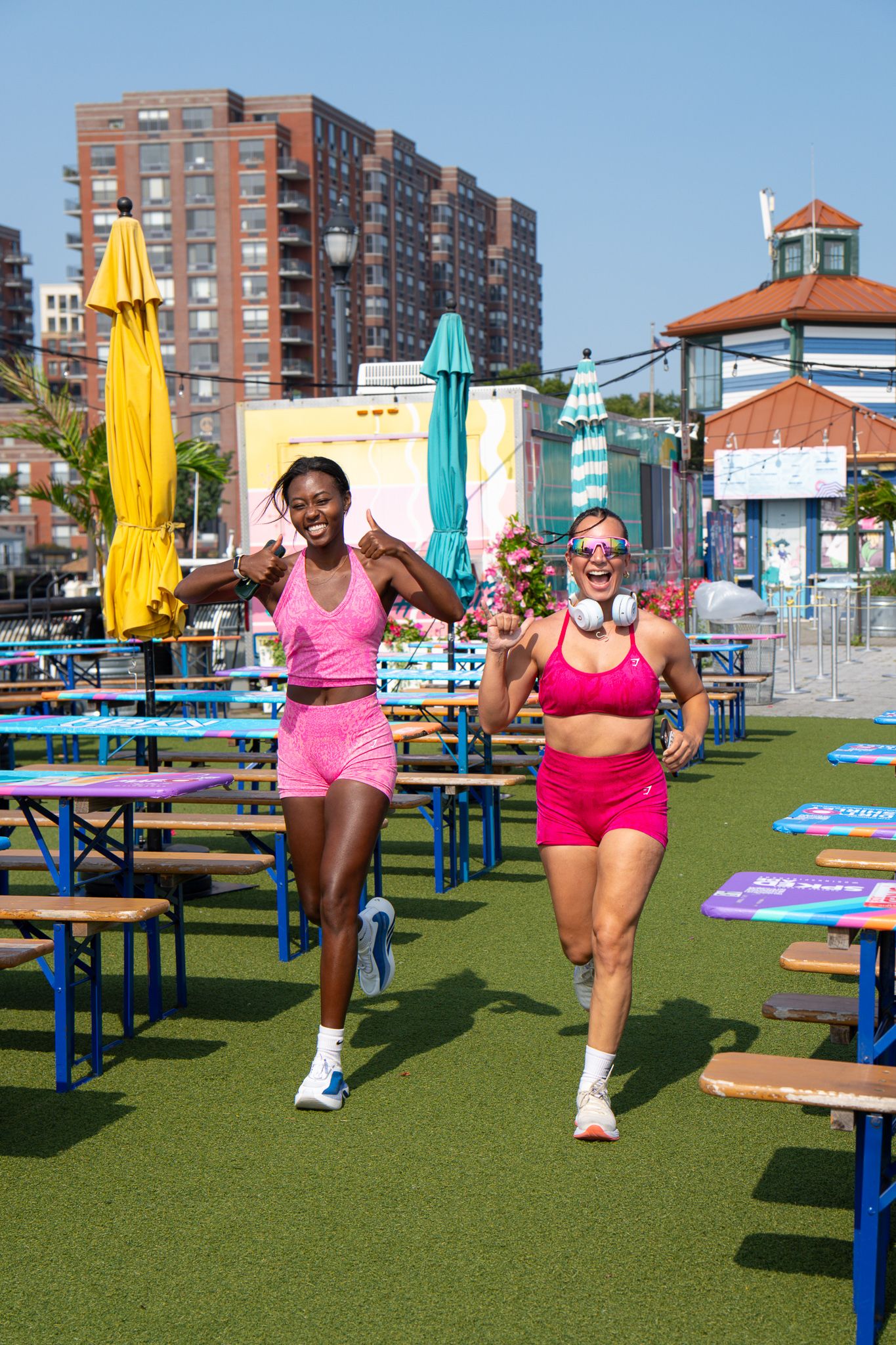 Two women in matching pink activewear run together at an outdoor fitness event with city buildings behind them.