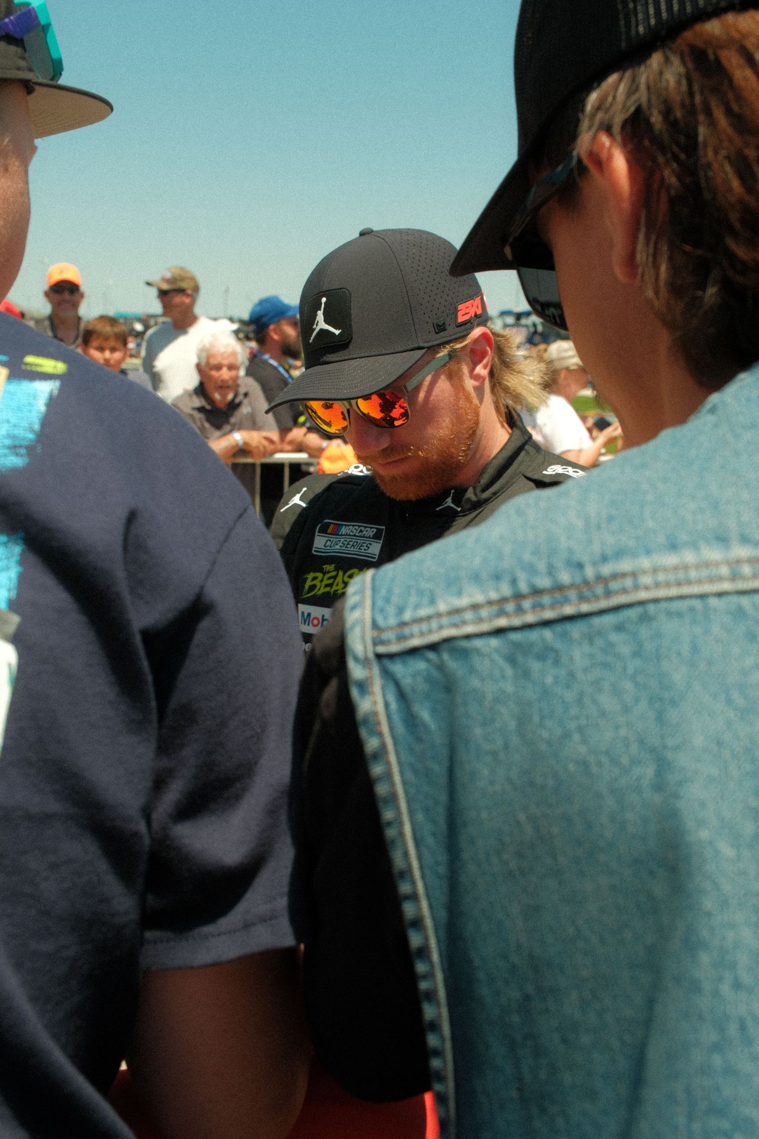 NASCAR driver signing autographs for fans at a sunny outdoor racing event, wearing a black Jordan cap and reflective sunglasses.