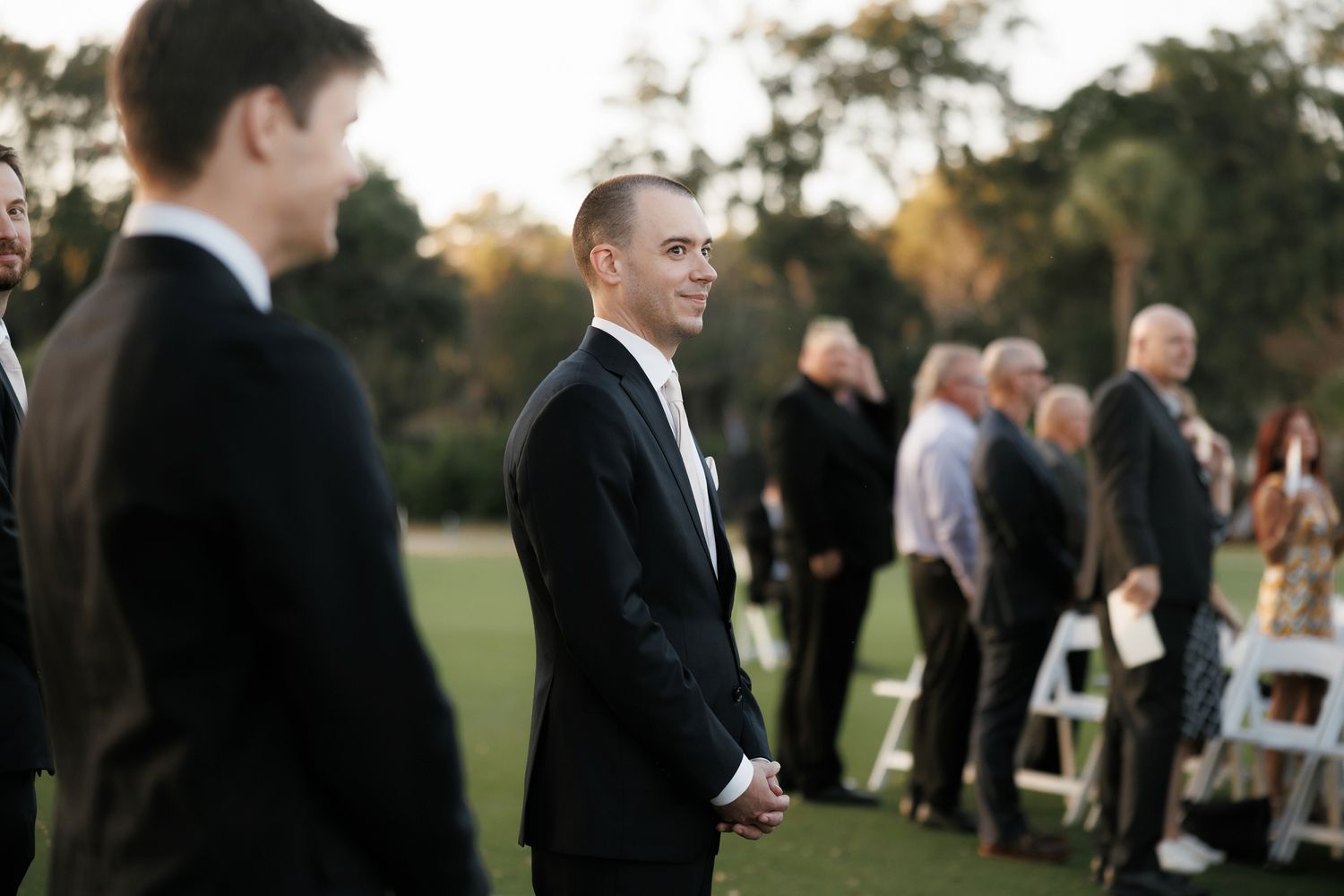 Wedding guests mingle and socialize outdoors during a golden sunset reception on a grassy lawn.
