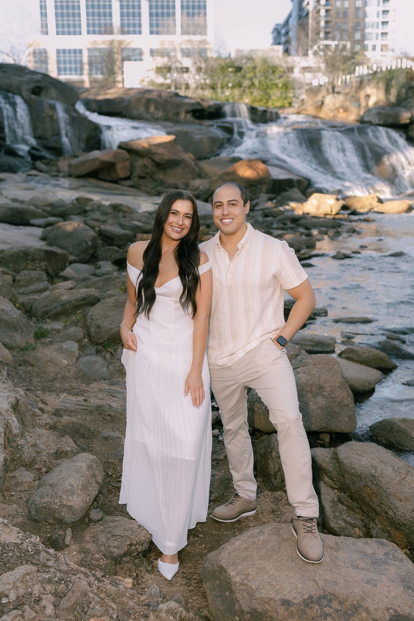 Couple in white clothing sitting together on rocky coastal terrain with buildings in background.