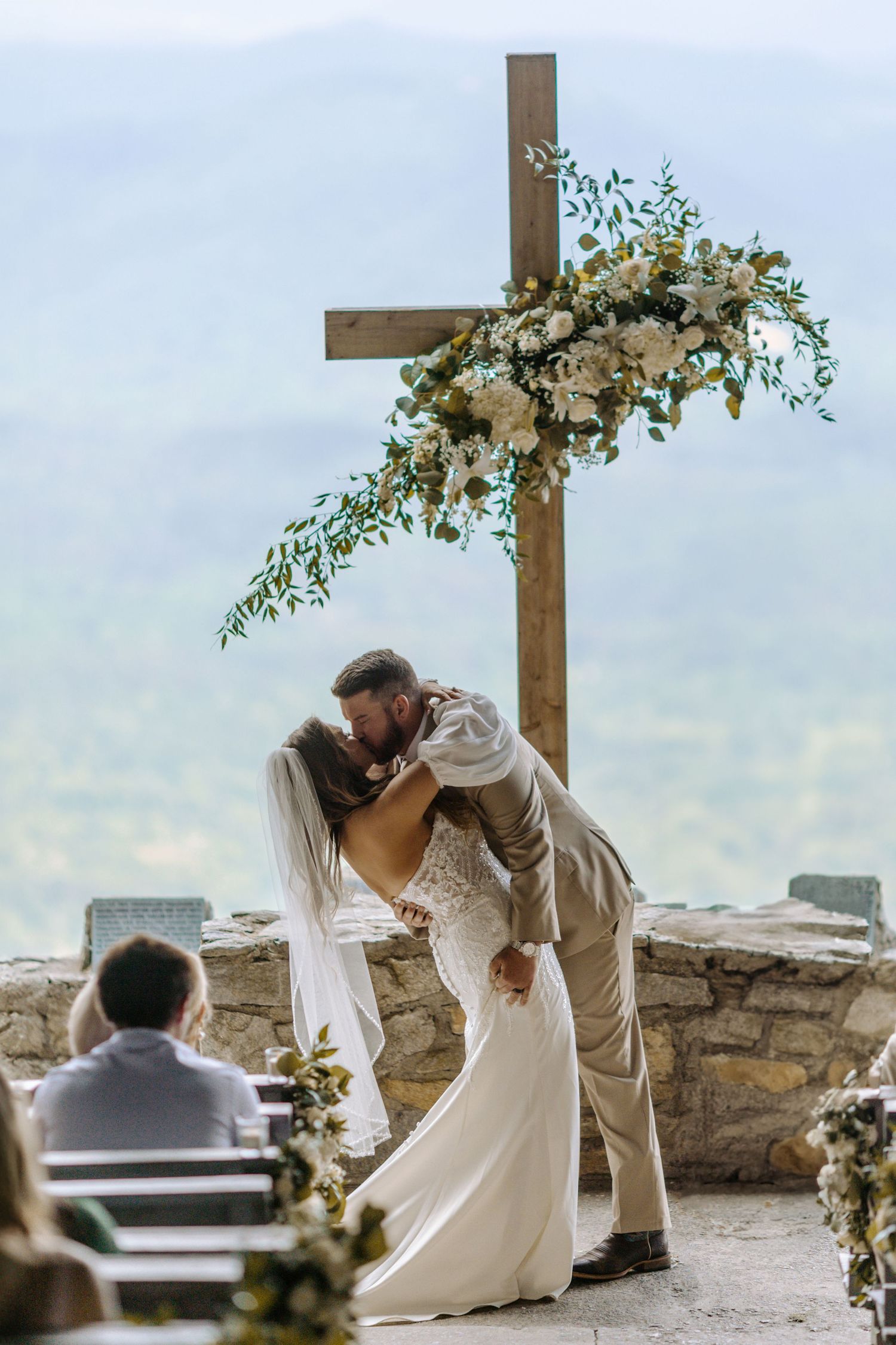 A romantic outdoor wedding ceremony with a wooden cross adorned with white flowers and greenery against a scenic mountain view.