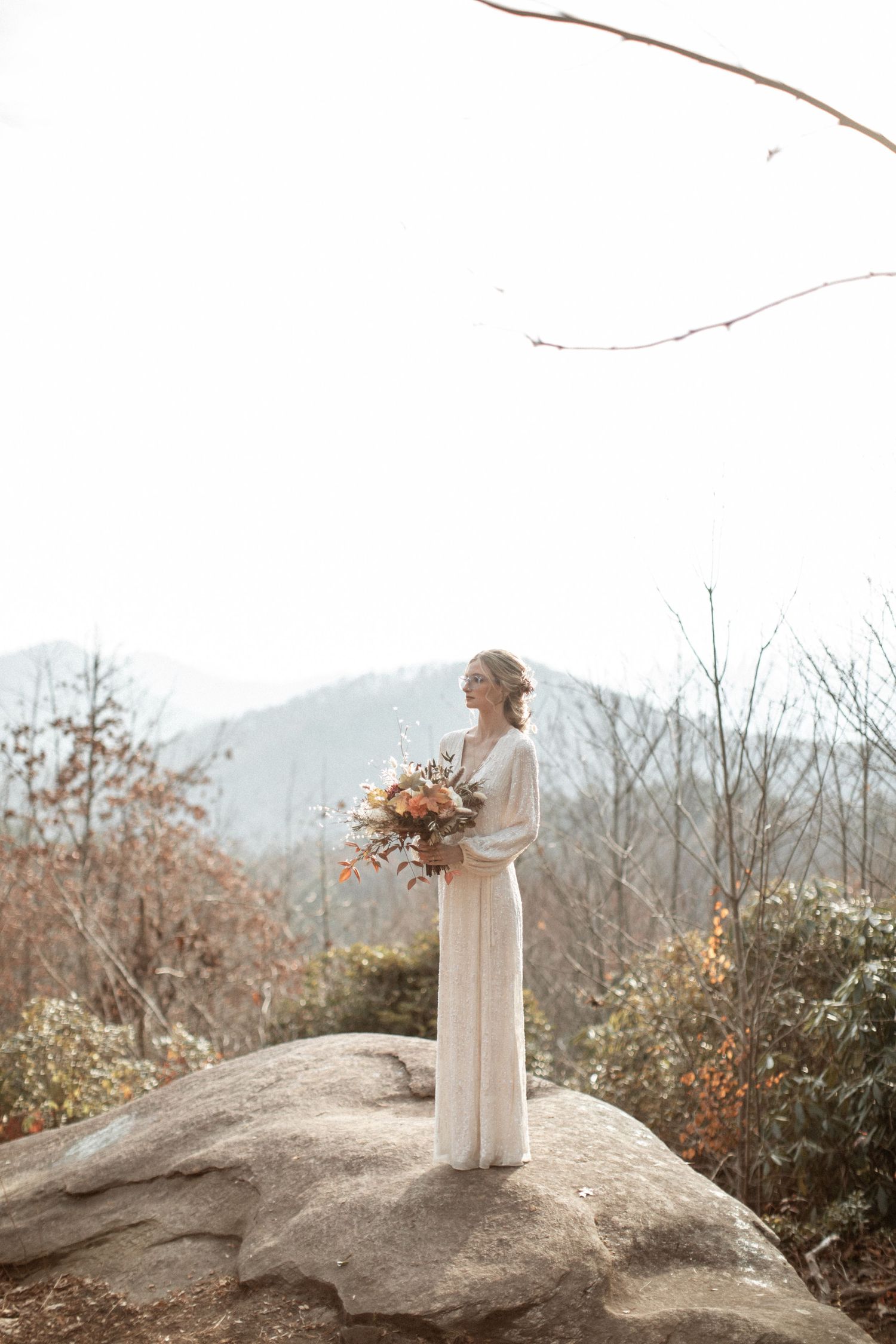 A bride in a flowing white dress stands on a rocky outcrop overlooking misty mountains.