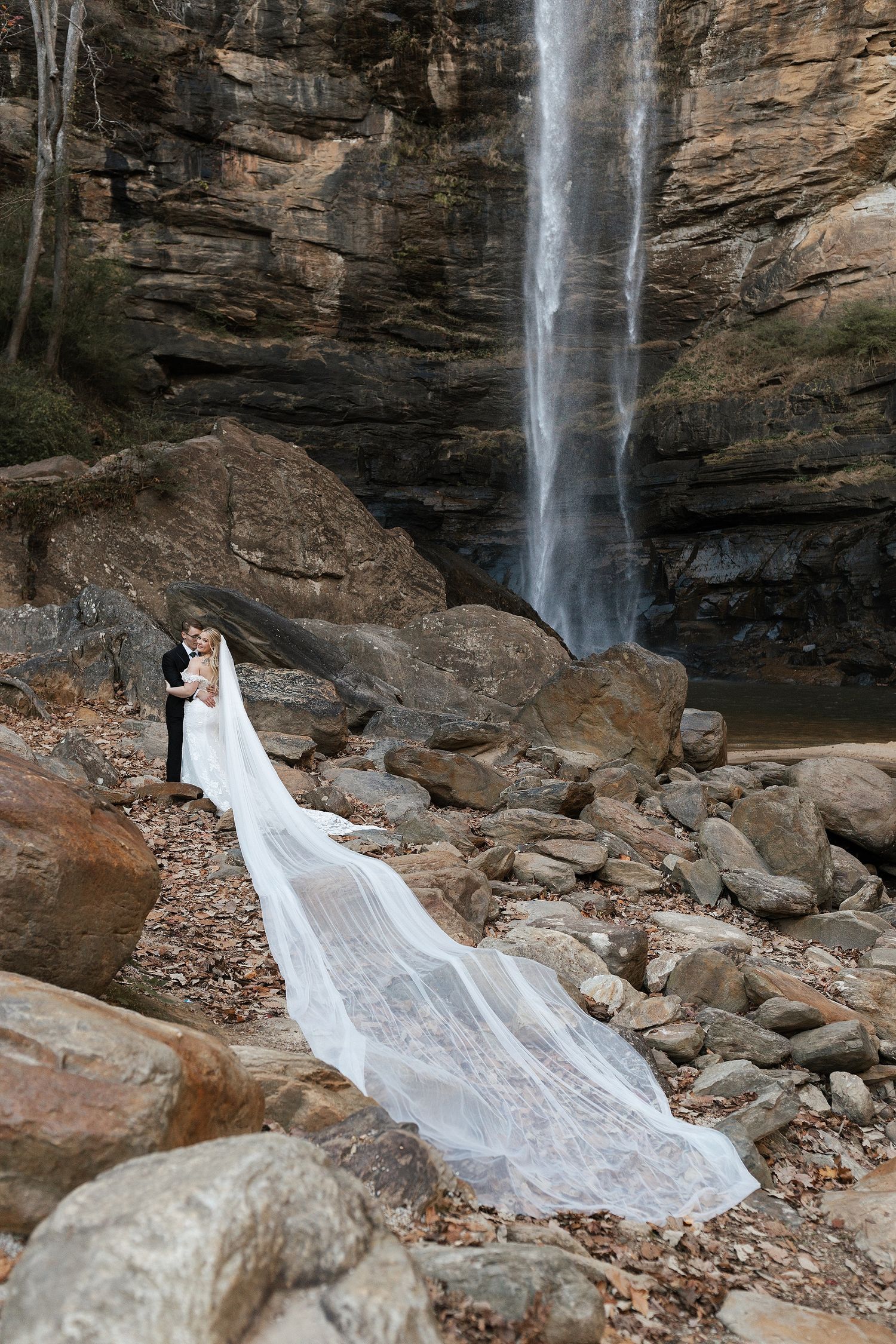 A scenic waterfall (Toccoa Falls) cascades down rocky cliffs as a flowing white dress train extends dramatically across the rocky terrain.