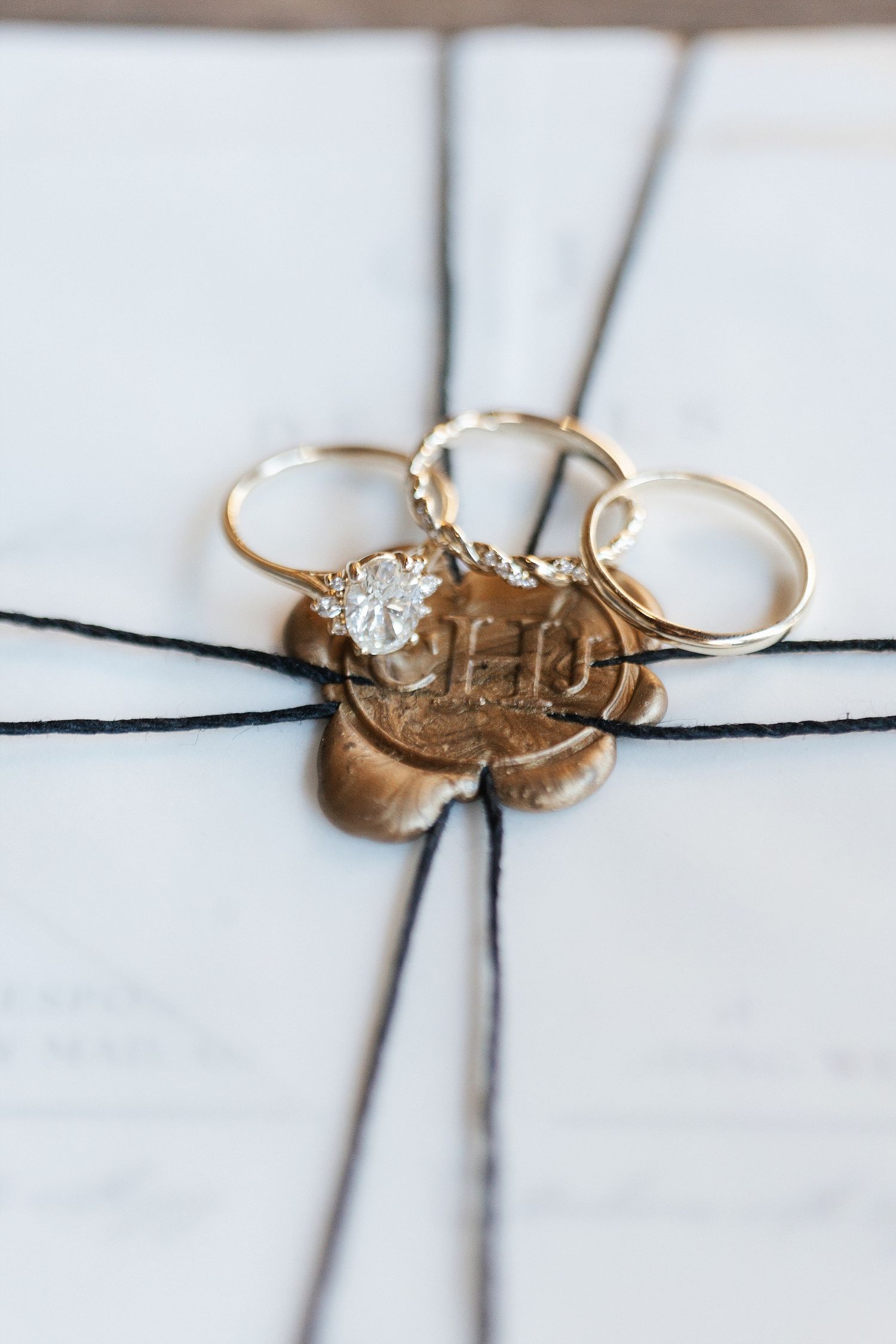 Wedding rings and diamond engagement ring displayed on gold wax seal tied with black string against white background.