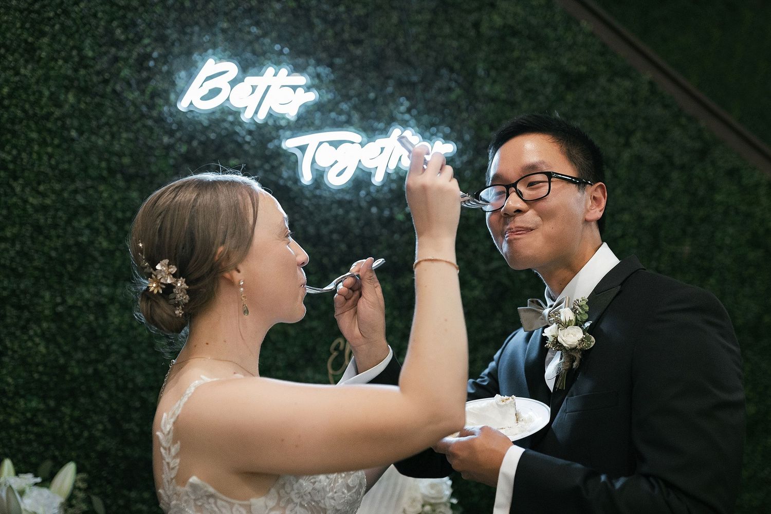 A couple feeds each other dessert in front of a blue neon 'Better Together' sign at their wedding celebration.