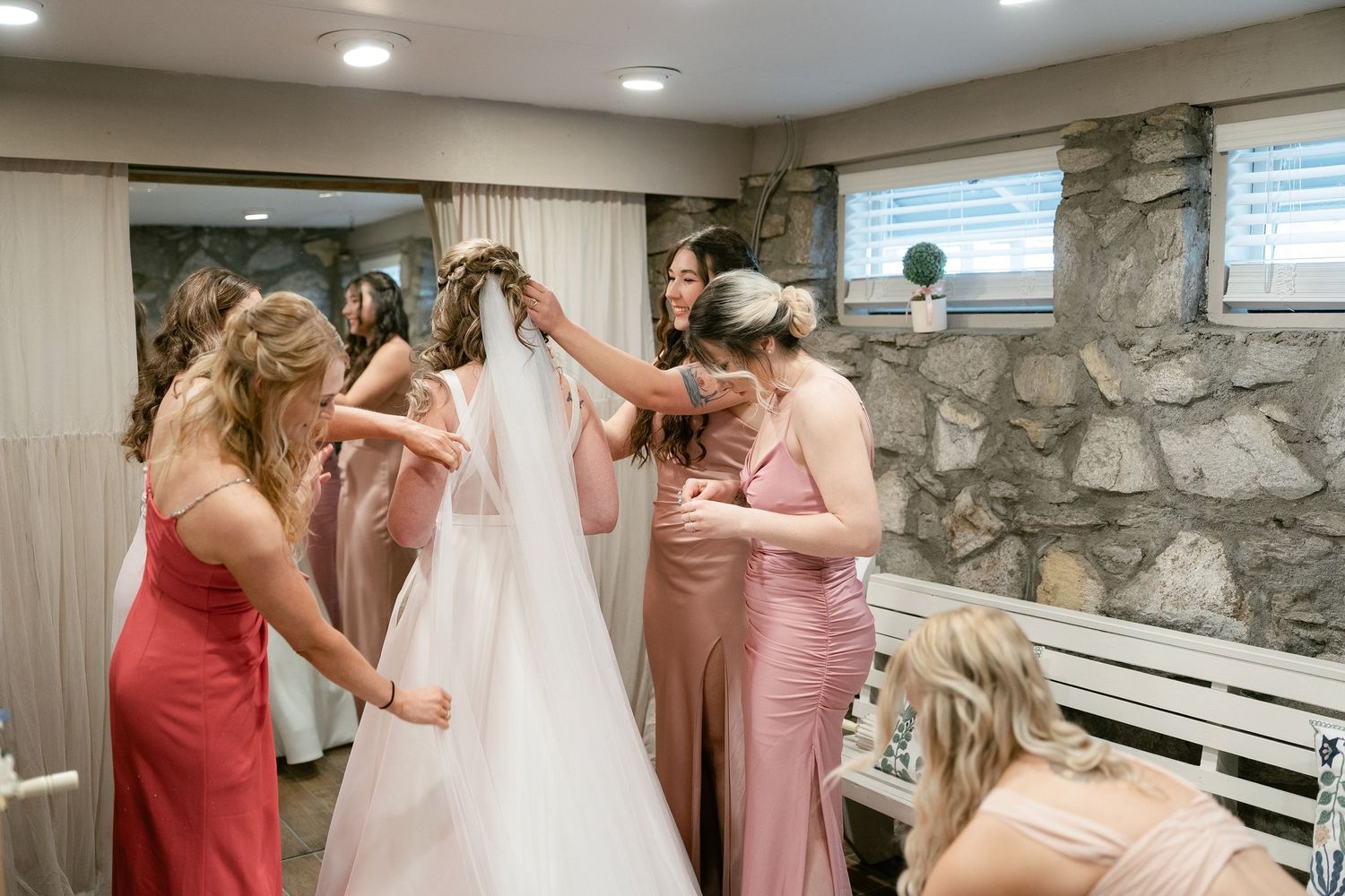 Wedding party helps bride get dressed in pink and white formal attire in stone-walled room with natural lighting.