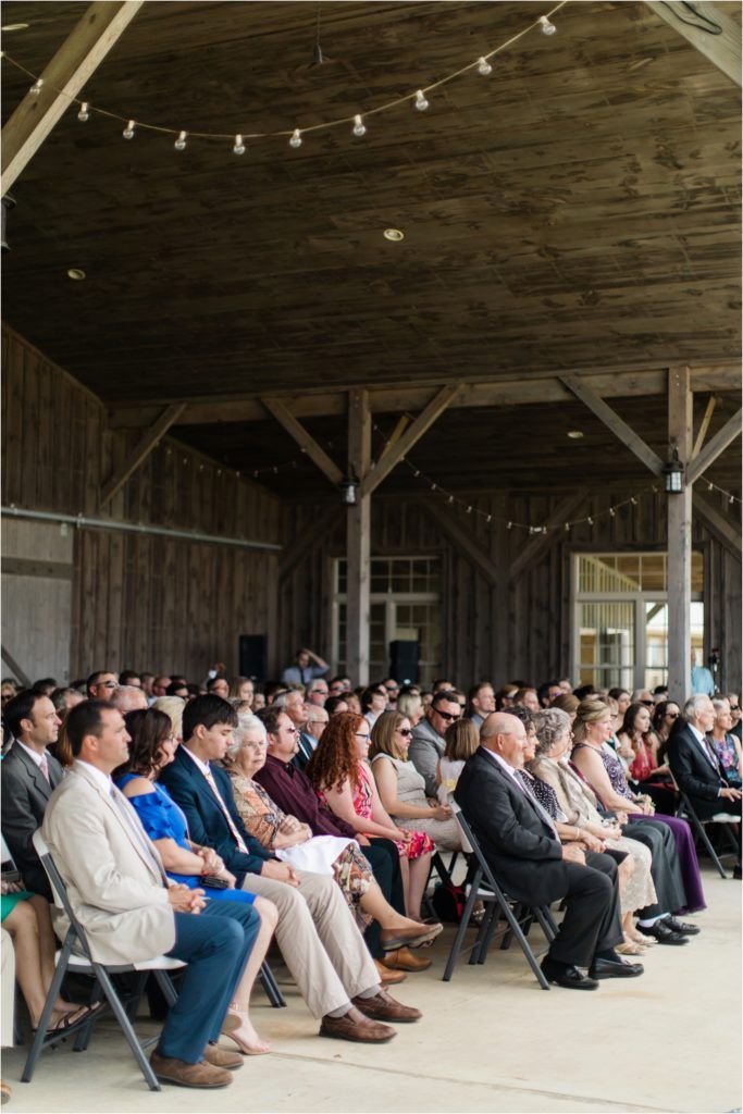Wedding guests seated in rustic barn venue with string lights overhead during ceremony.