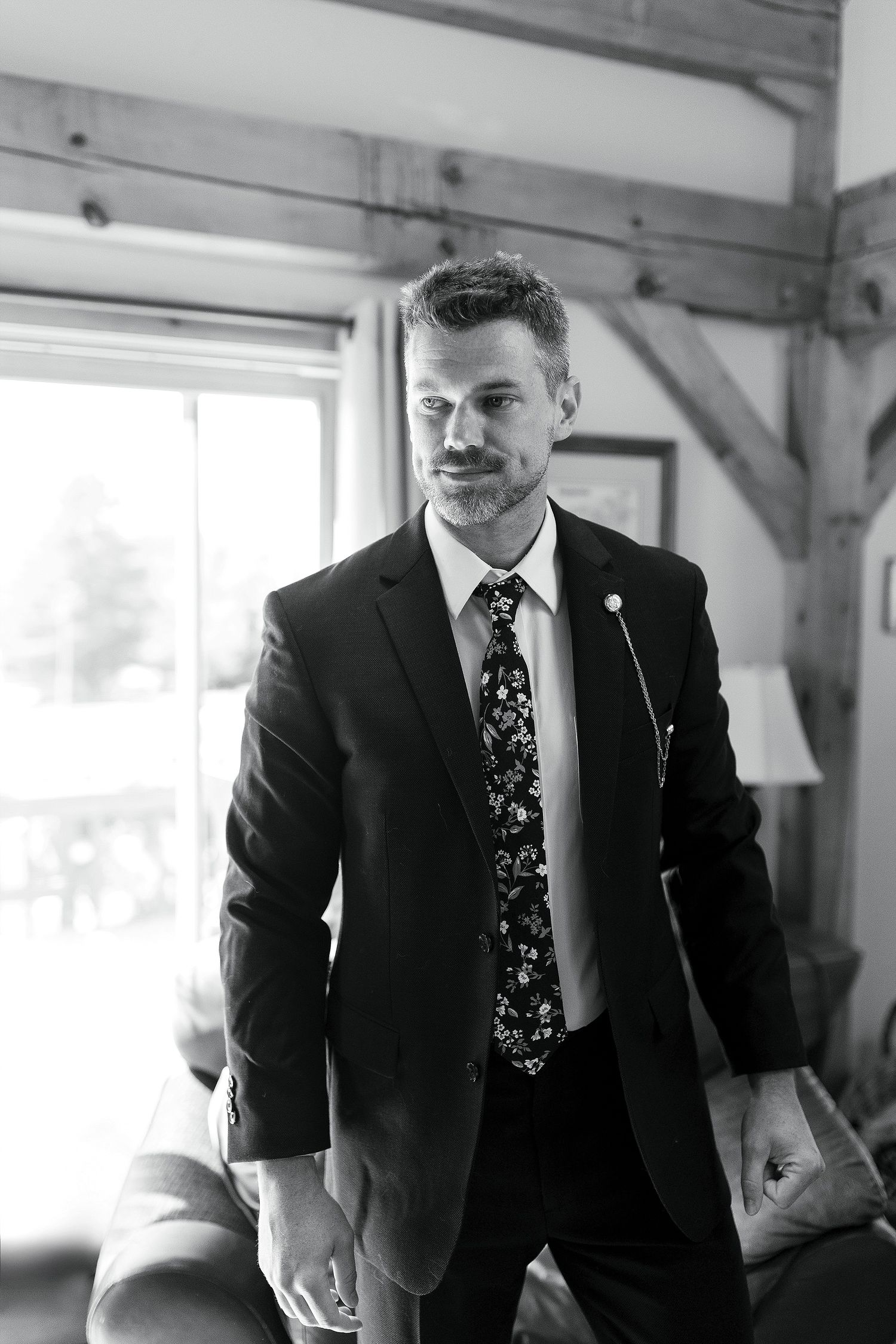Man in dark suit and floral tie standing in rustic indoor setting.