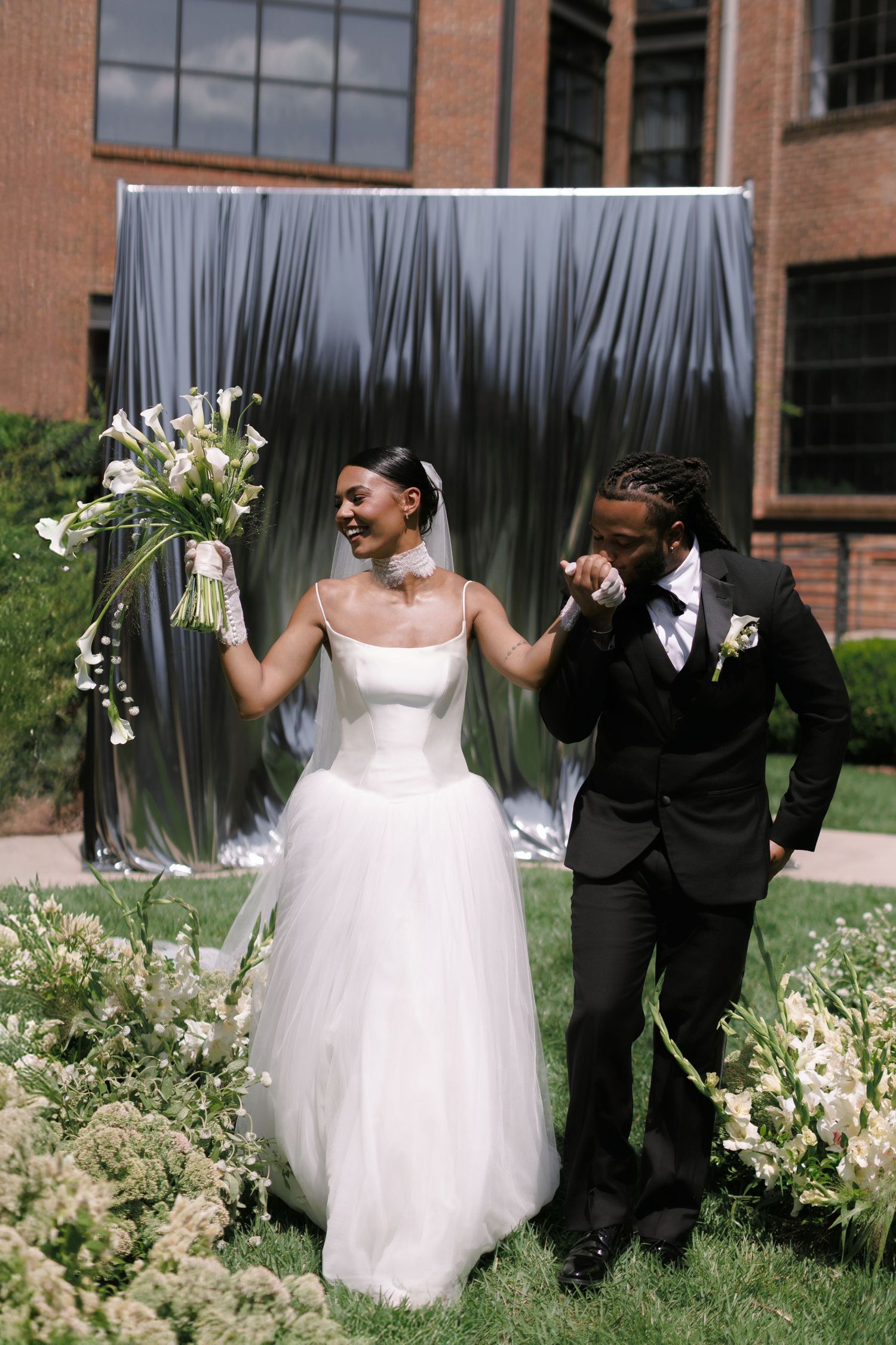 Bride in white tulle gown holds bouquet while groom in black suit kisses her hand outdoors at elegant wedding ceremony.