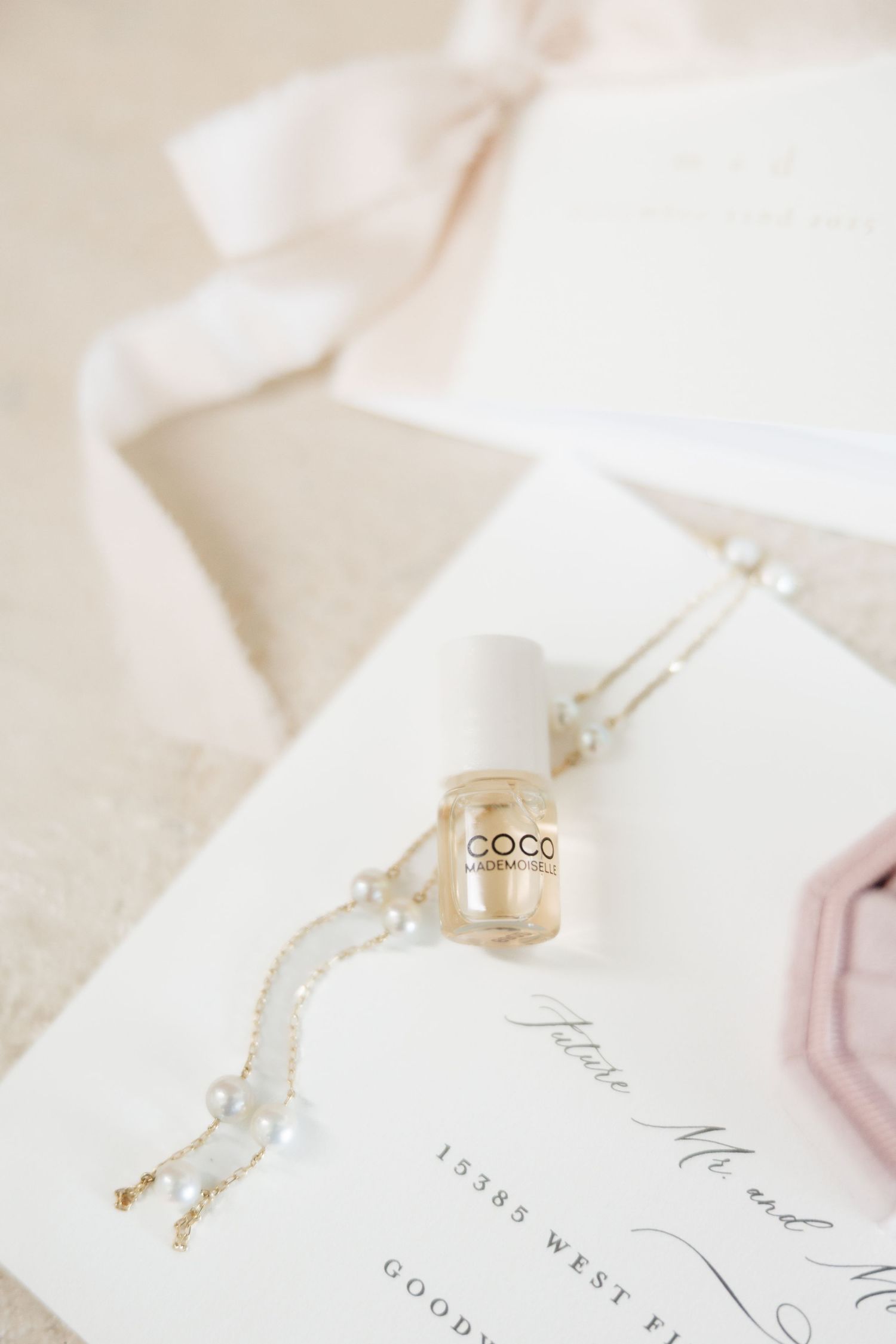Elegant wedding flat lay with COCO nail polish, pearl bracelet, pink ribbon, and calligraphy invitation on white background.