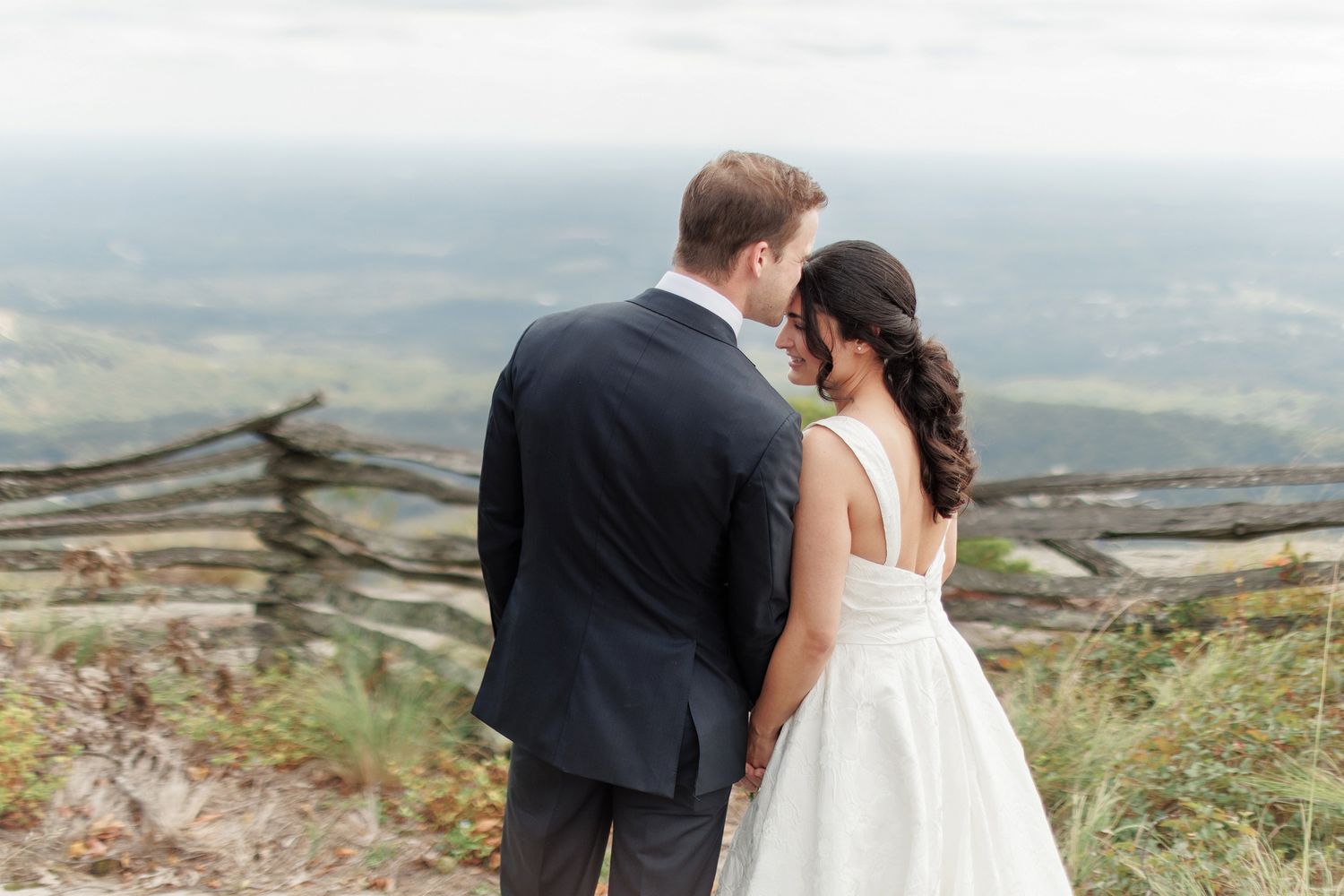 A bride and groom stand together on a mountain overlook, sharing an intimate moment with scenic valley views behind them.