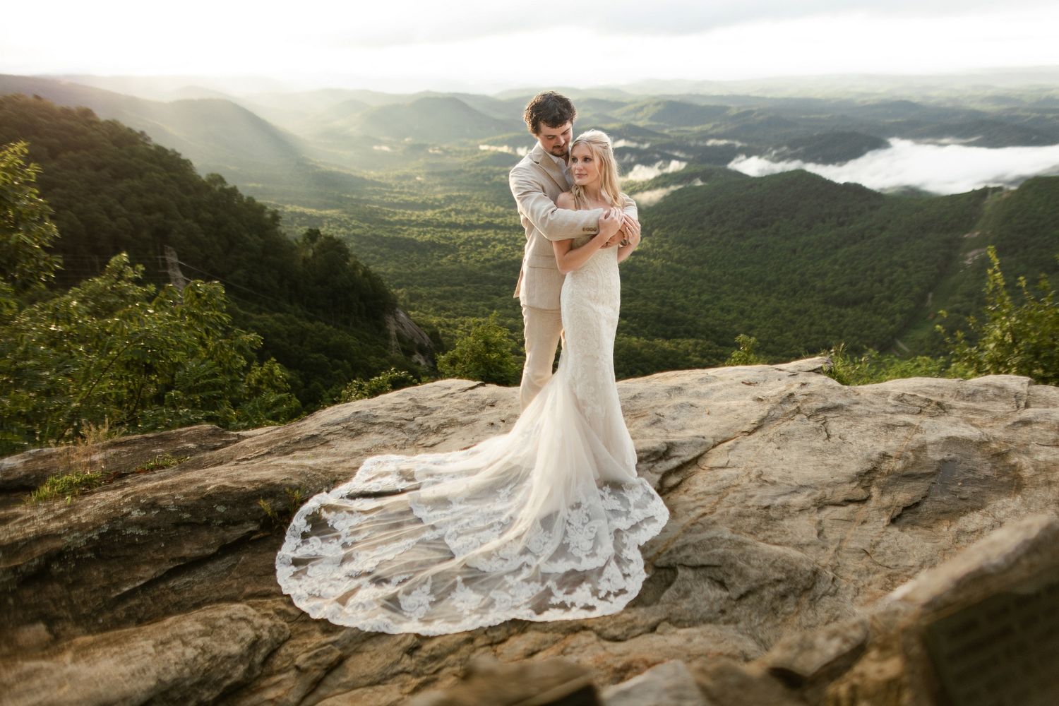 Bride and groom embracing on rocky mountain summit, bride in lace gown with long train at sunrise.