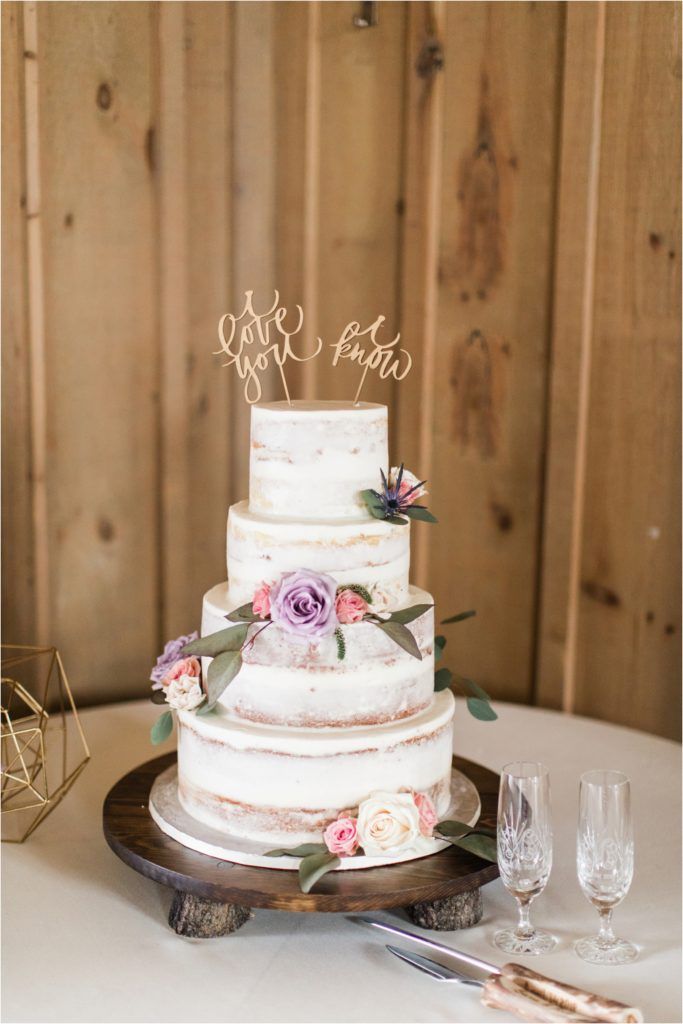 Three-tiered semi-naked wedding cake decorated with pink and purple roses on a rustic wooden stand.