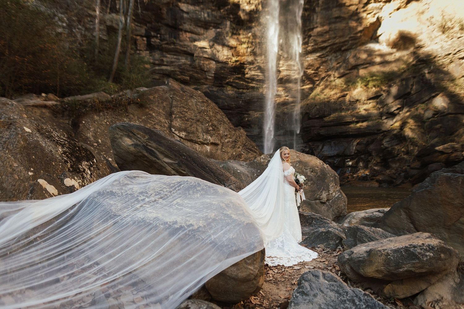 A flowing white wedding dress veil catches the wind dramatically beneath a towering natural waterfall and rocky cliffs.