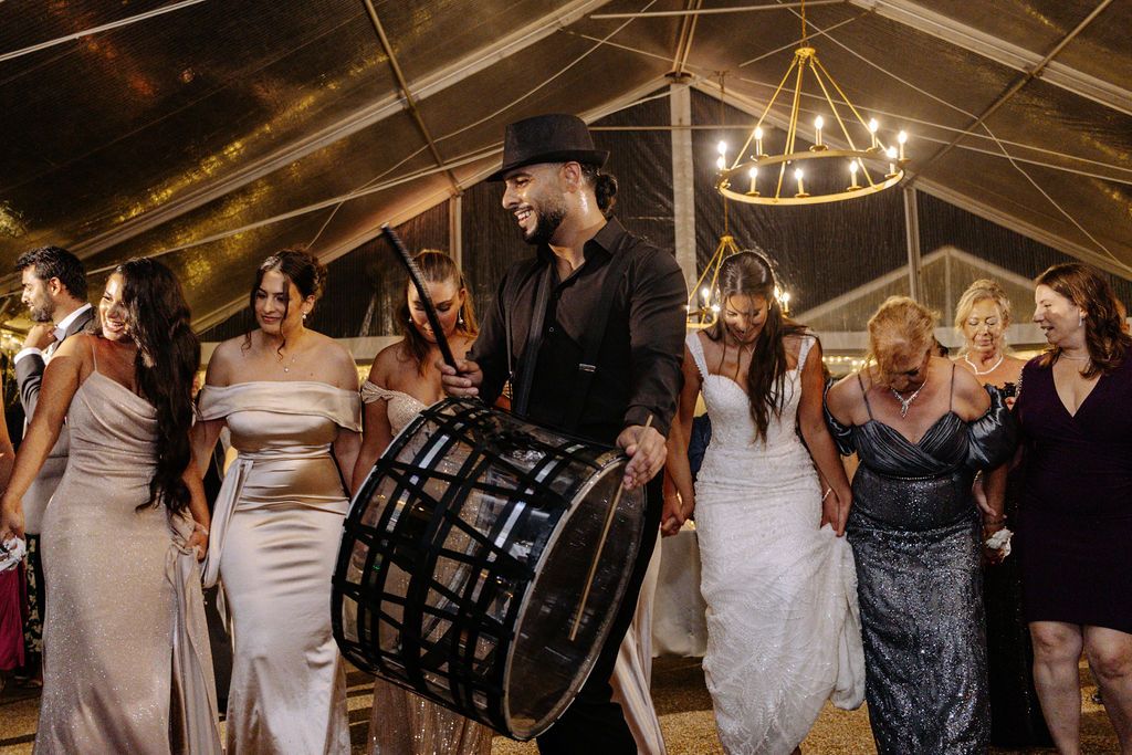 Wedding party dancing around a drummer during a lively reception celebration under a tent.