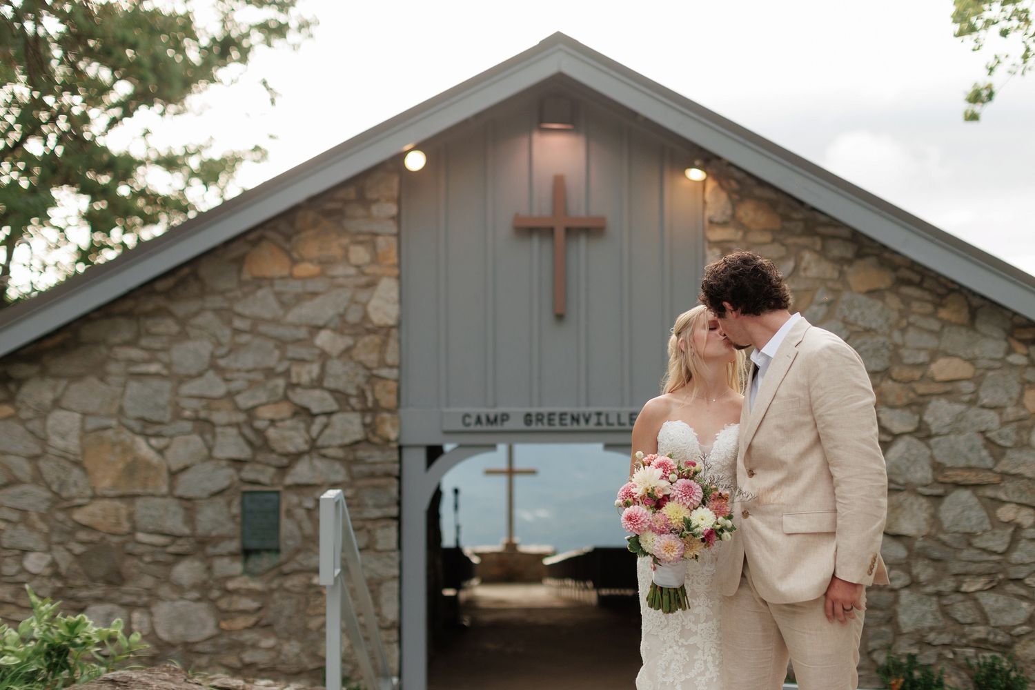 A couple shares an intimate moment outside a rustic stone chapel with a wooden cross on the facade.