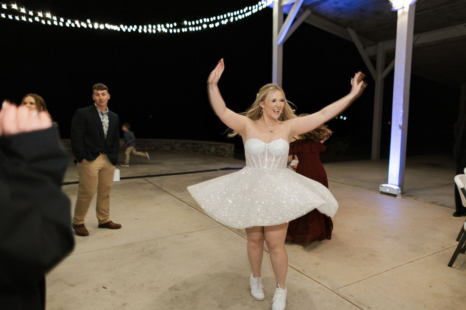 A dancer in a white dress and sneakers twirls on a dance floor under string lights at an outdoor event.