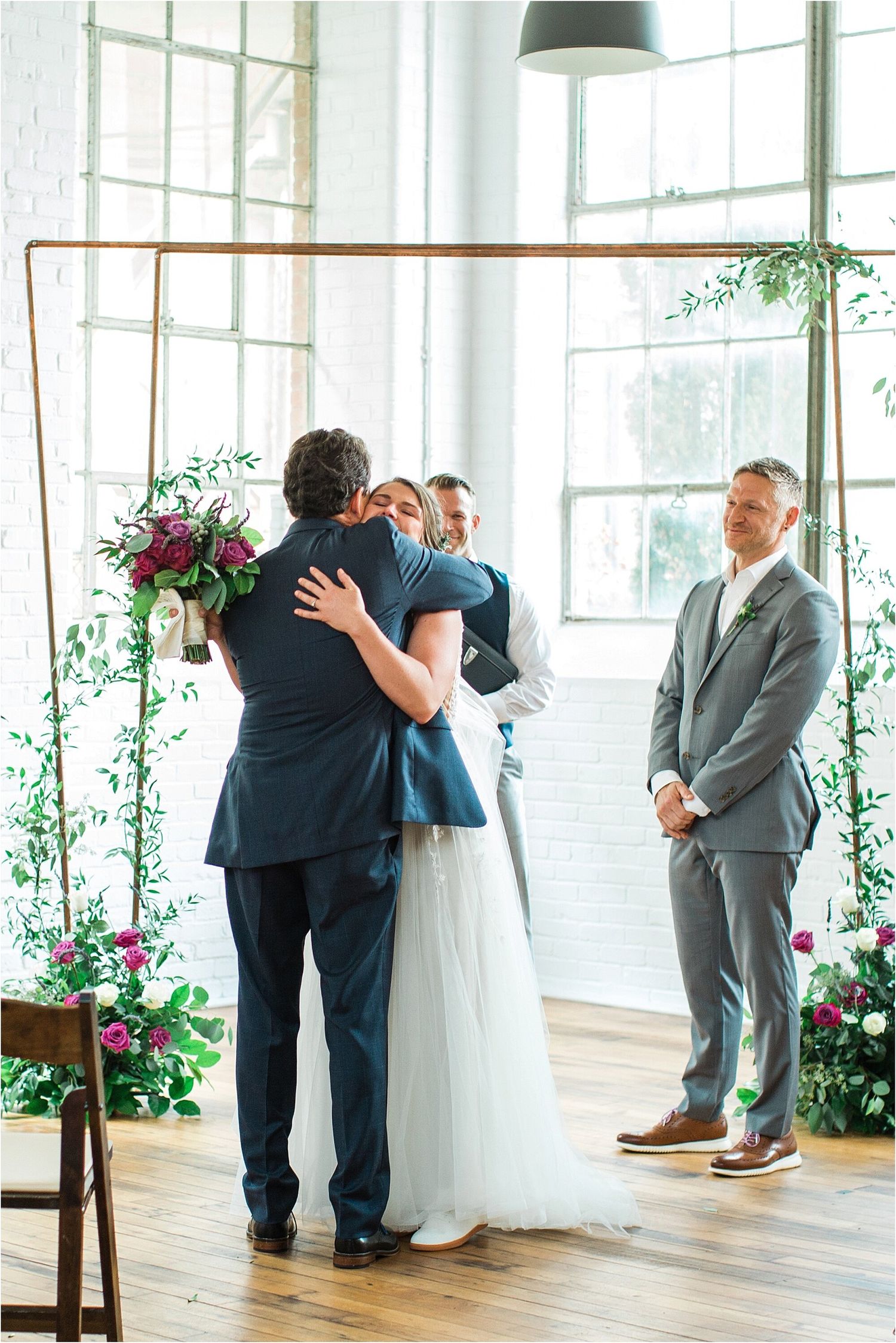 Emotional embrace during wedding ceremony in bright industrial space with copper arch and floral decor.