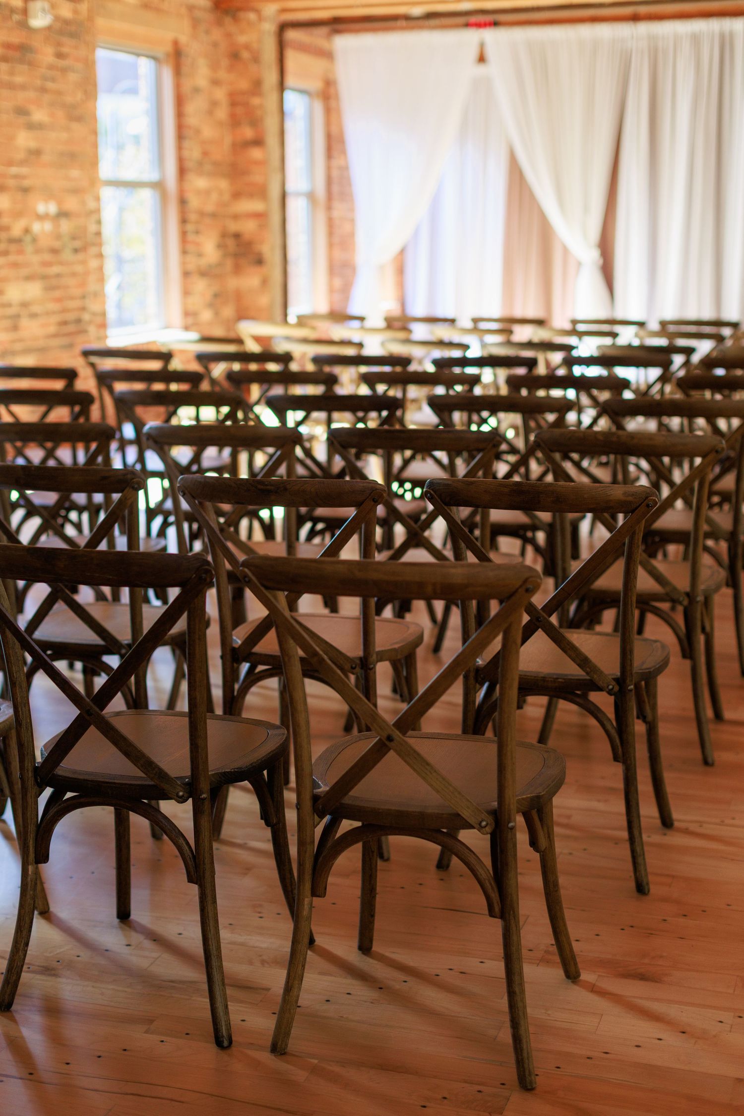 Rows of rustic wooden cross-back chairs arranged for a wedding ceremony in a brick-walled venue with white drape backdrop.