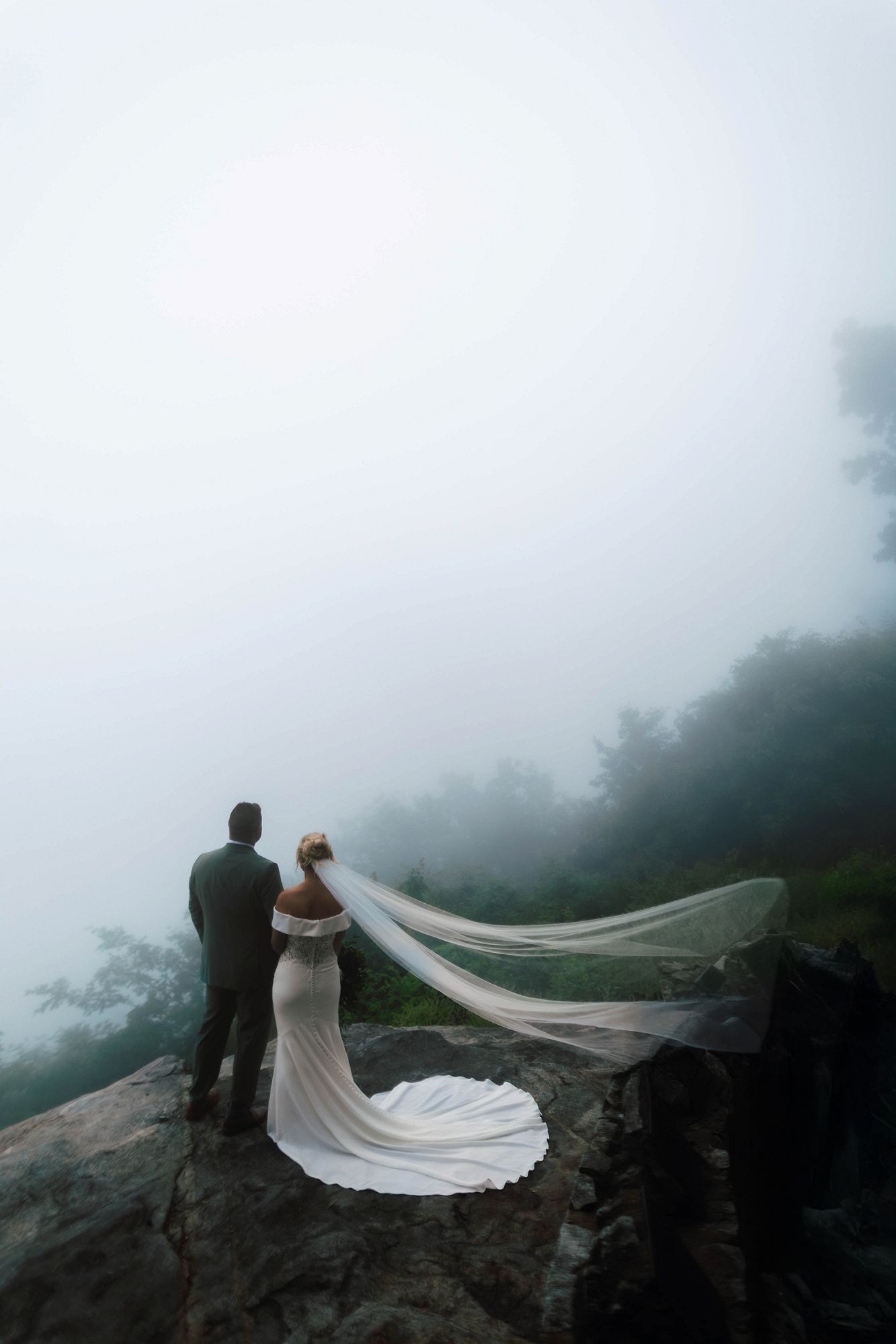 Bride and groom standing on a rocky cliff edge, bride's veil billowing in wind against a misty landscape.