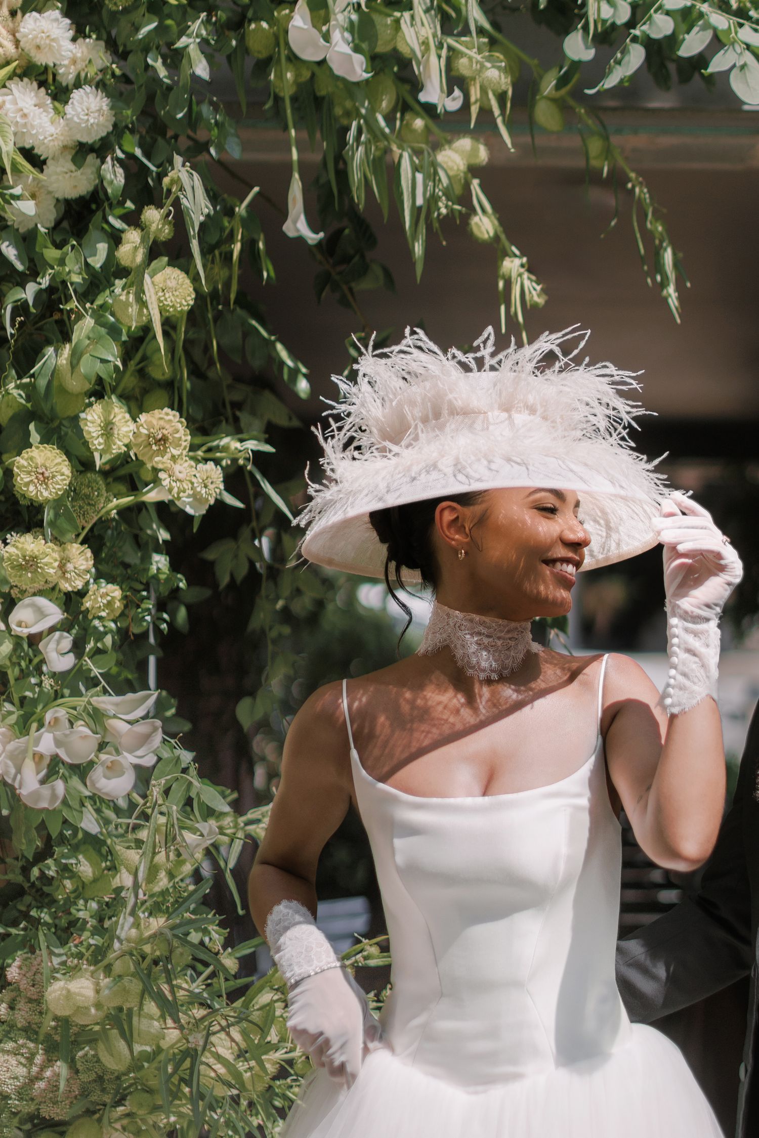 Smiling bride in white satin gown and feathered wide-brim hat poses among lush white floral arrangements.