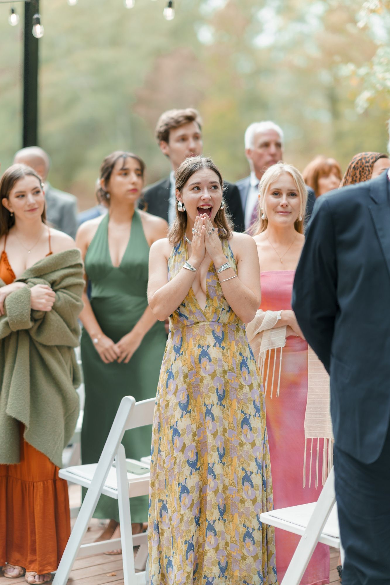 Emotional wedding guest in yellow floral dress reacts with joy during outdoor ceremony surrounded by guests.